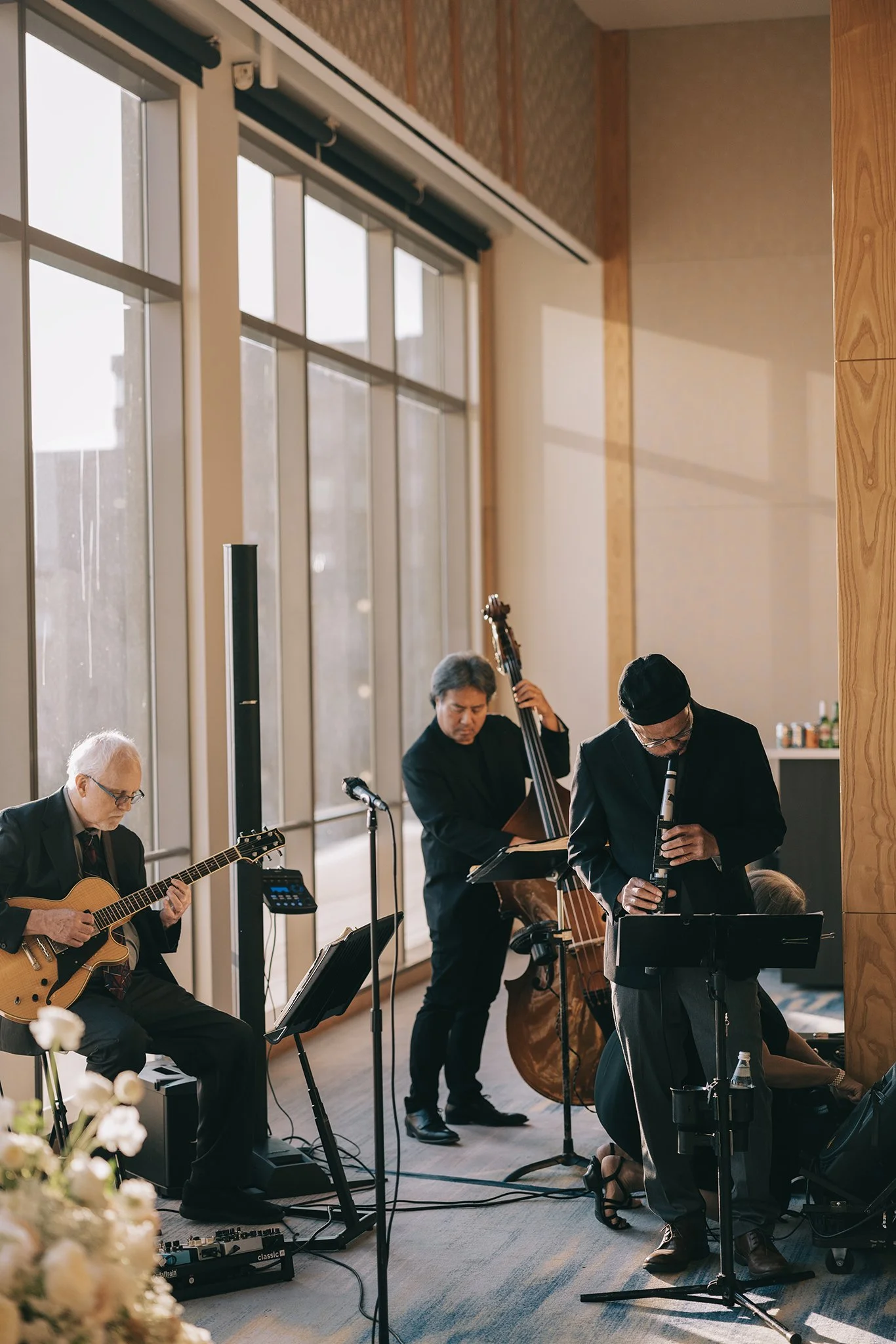 A jazz band performing indoors near large windows with sunlight streaming in, featuring guitar, double bass, and wind instruments.