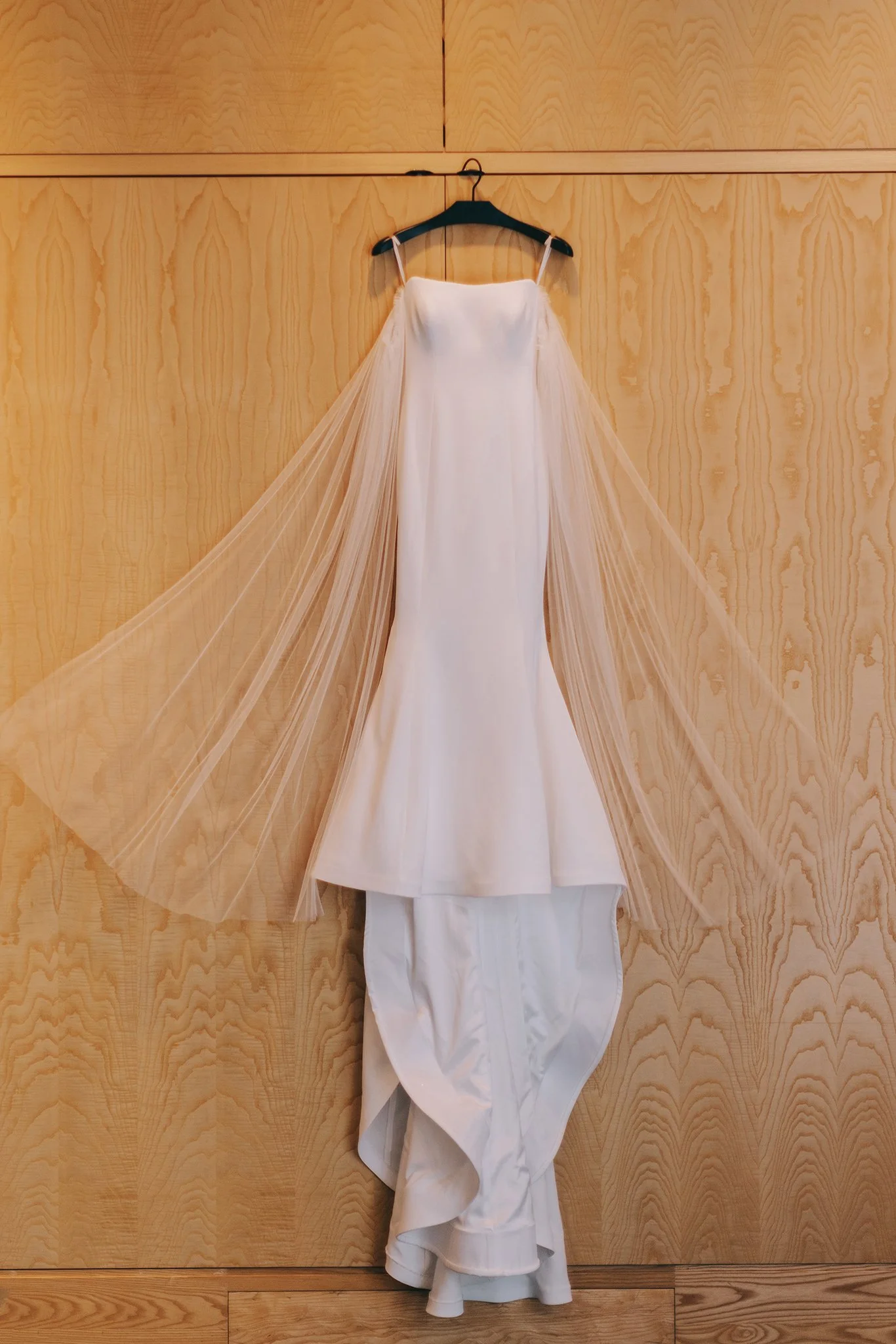 A wedding dress hanging on a black hanger against a wooden wall, with a fitted bodice, thin straps, and a long train of tulle and satin.
