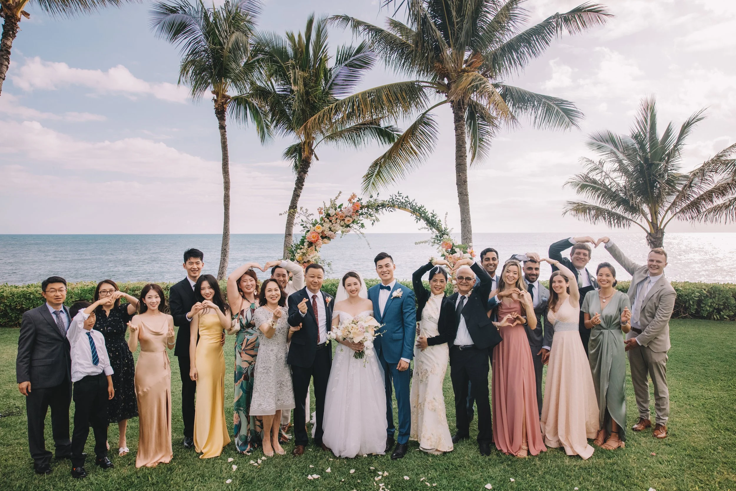 A group of people at a wedding, standing outdoors on grass near the ocean, with palm trees and a floral arch in the background.