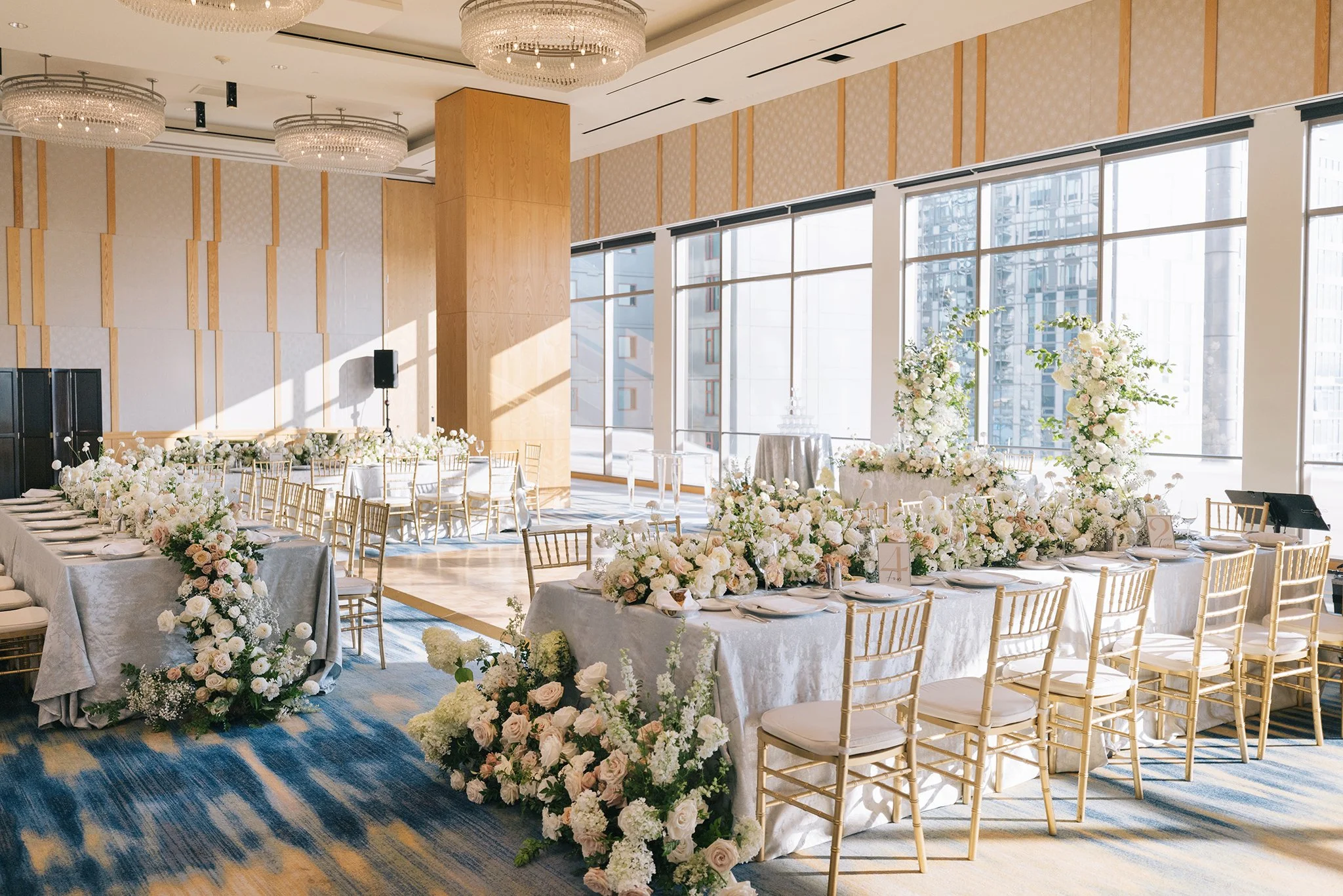 Elegant banquet hall decorated with white flowers, gold chairs, and long tables for a wedding or event, with large windows and city views.