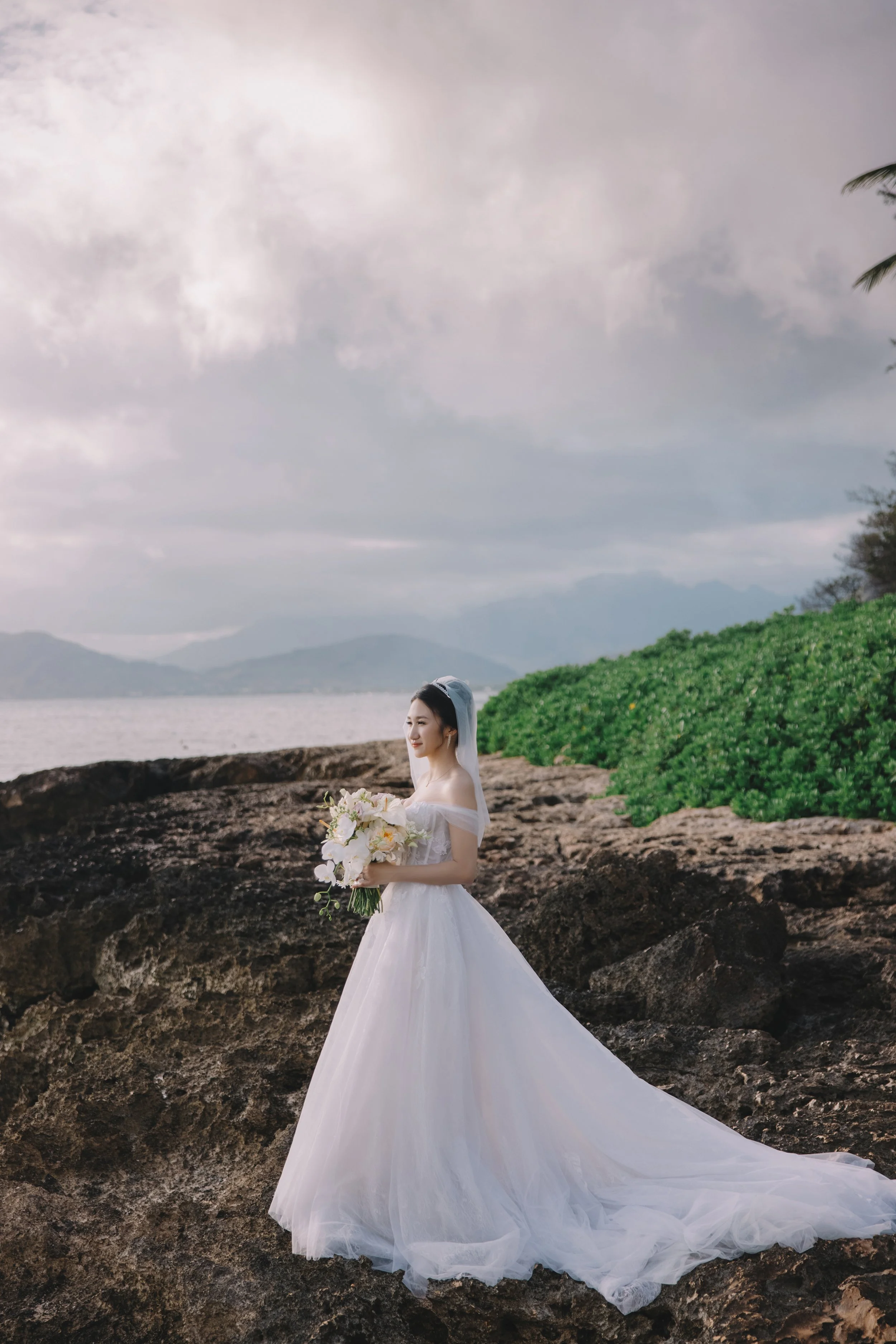 A bride in a white wedding gown holding a bouquet of flowers standing on rocky terrain near water with mountains and cloudy sky in the background.