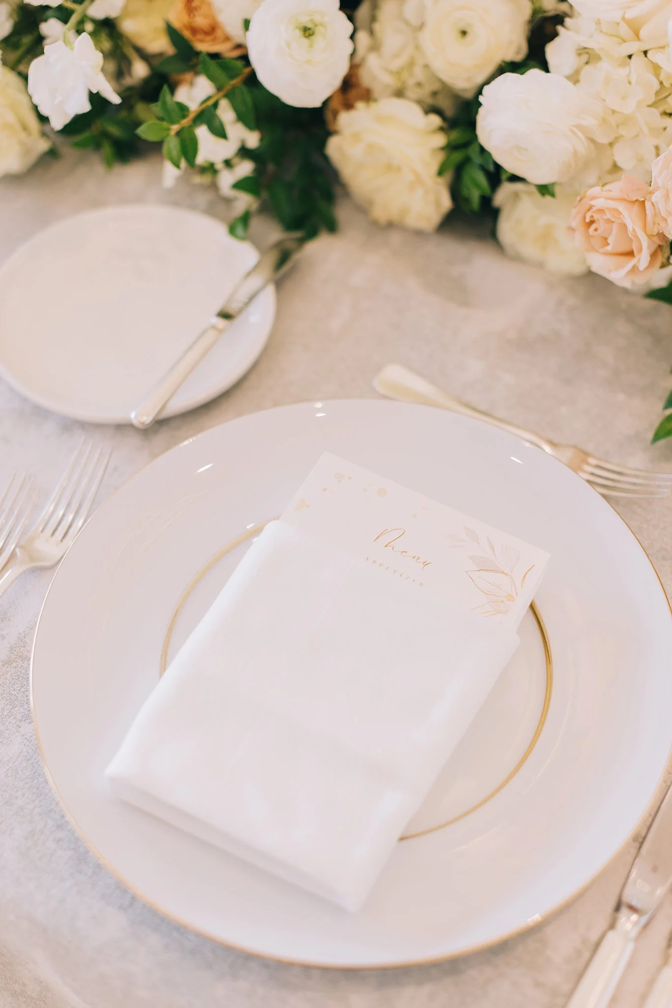 Elegant table setting with a white plate, gold rim, white napkin, and a menu card placed on top, surrounded by a floral centerpiece of white and blush pink roses.