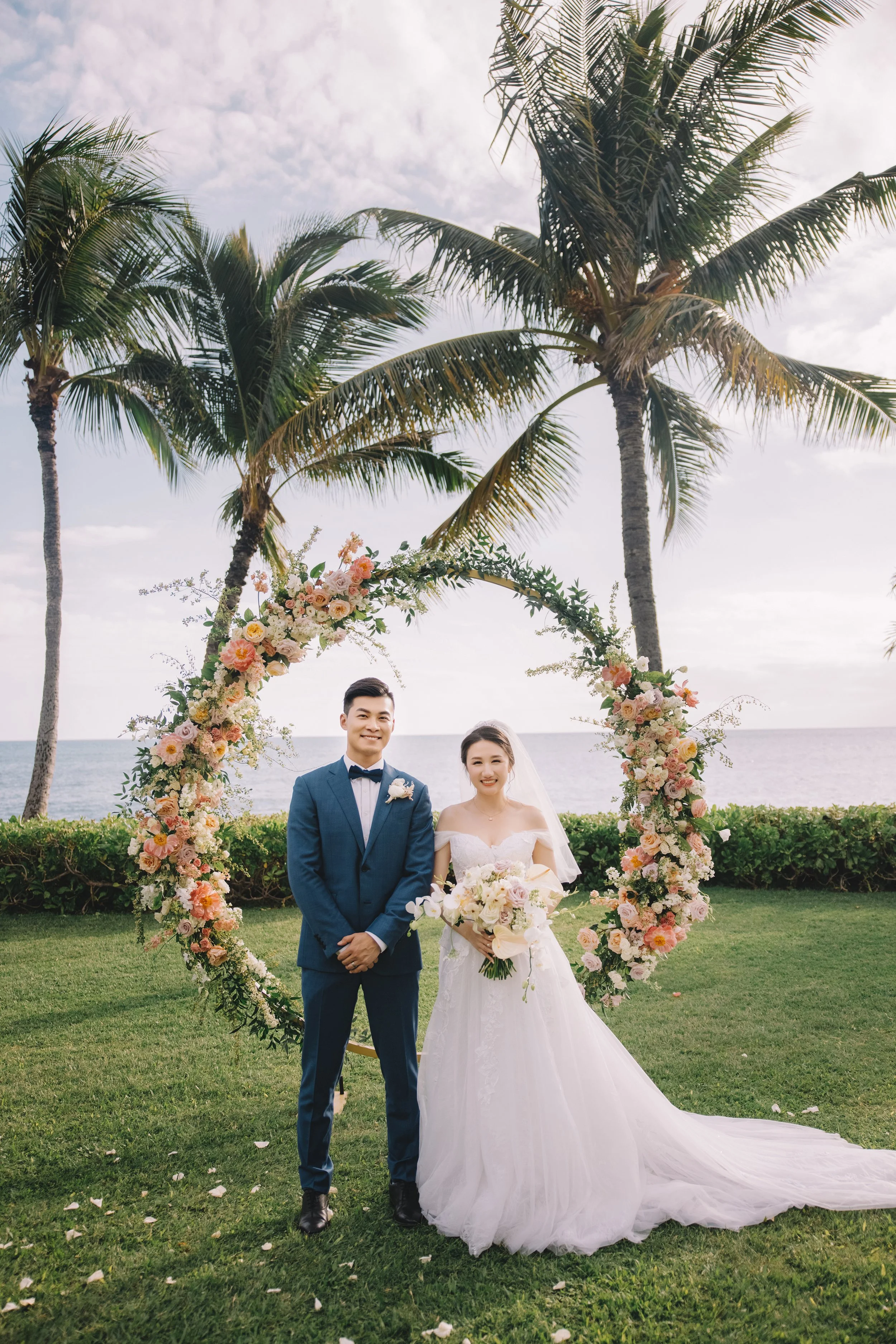 A newlywed couple standing in front of a floral ark on a lawn near the ocean, with palm trees and a cloudy sky in the background.