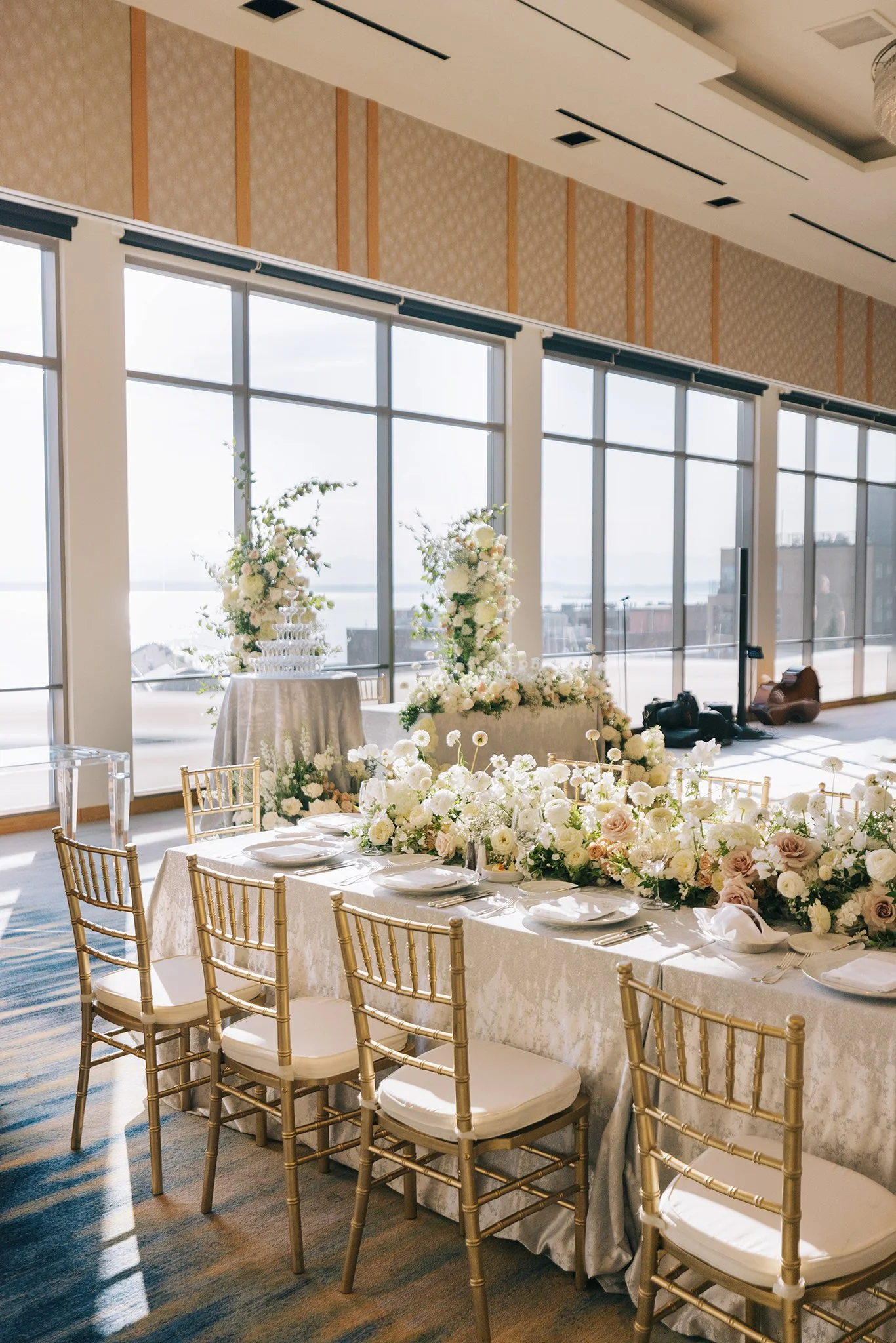 Elegant wedding reception setup with a long table decorated with white flowers and gold chairs, large windows in the background letting in natural light.