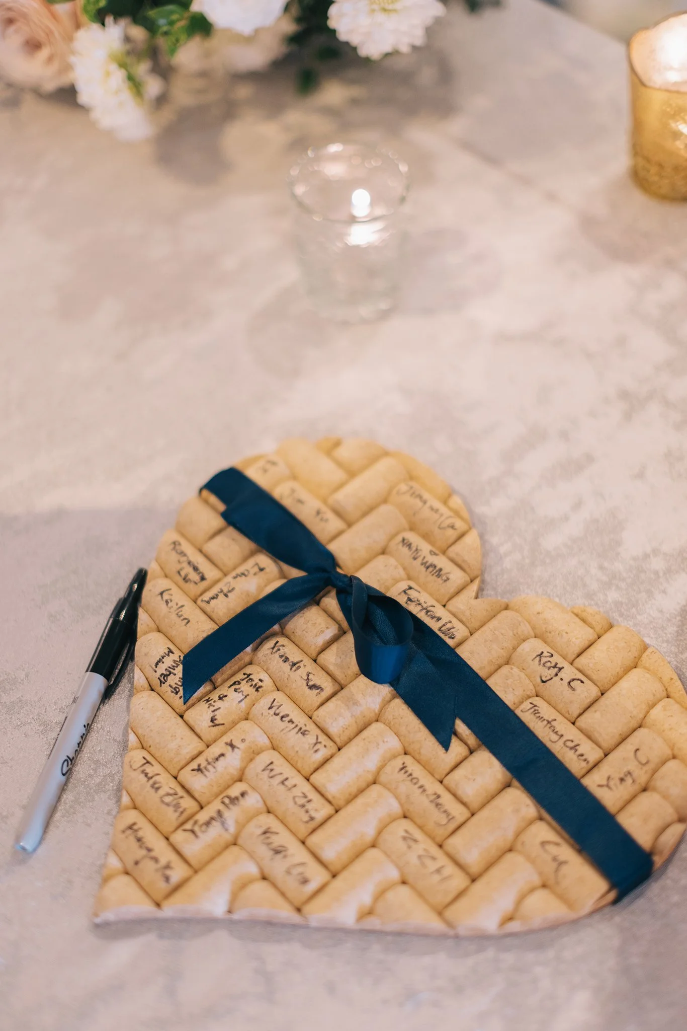 Heart-shaped arrangement of wine corks tied with a dark blue ribbon, placed on a table with a pen nearby.