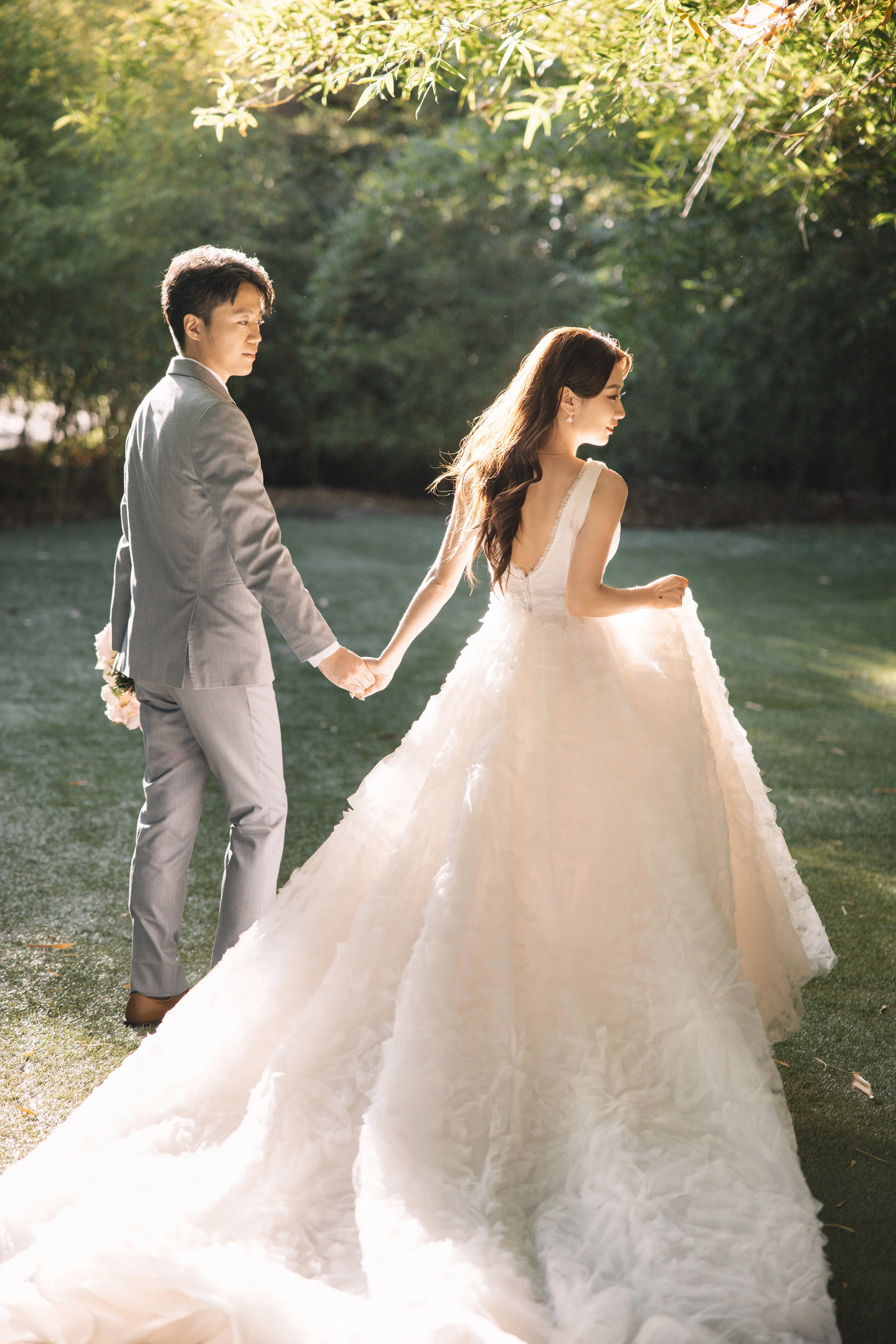 A couple dressed in wedding attire holding hands outdoors, with the bride in a voluminous white gown and the groom in a light gray suit, surrounded by greenery.