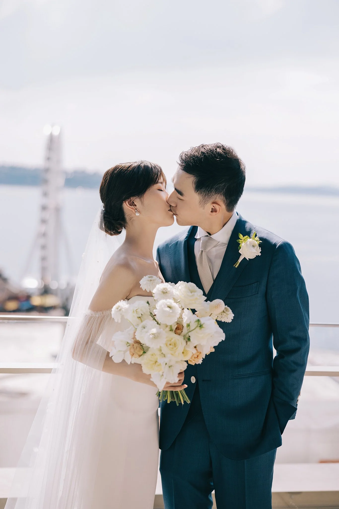 A bride in a white wedding dress holding a bouquet of white and cream flowers, kissing a groom in a blue suit with a boutonniere, by a body of water with an outdoor ferris wheel in the background.