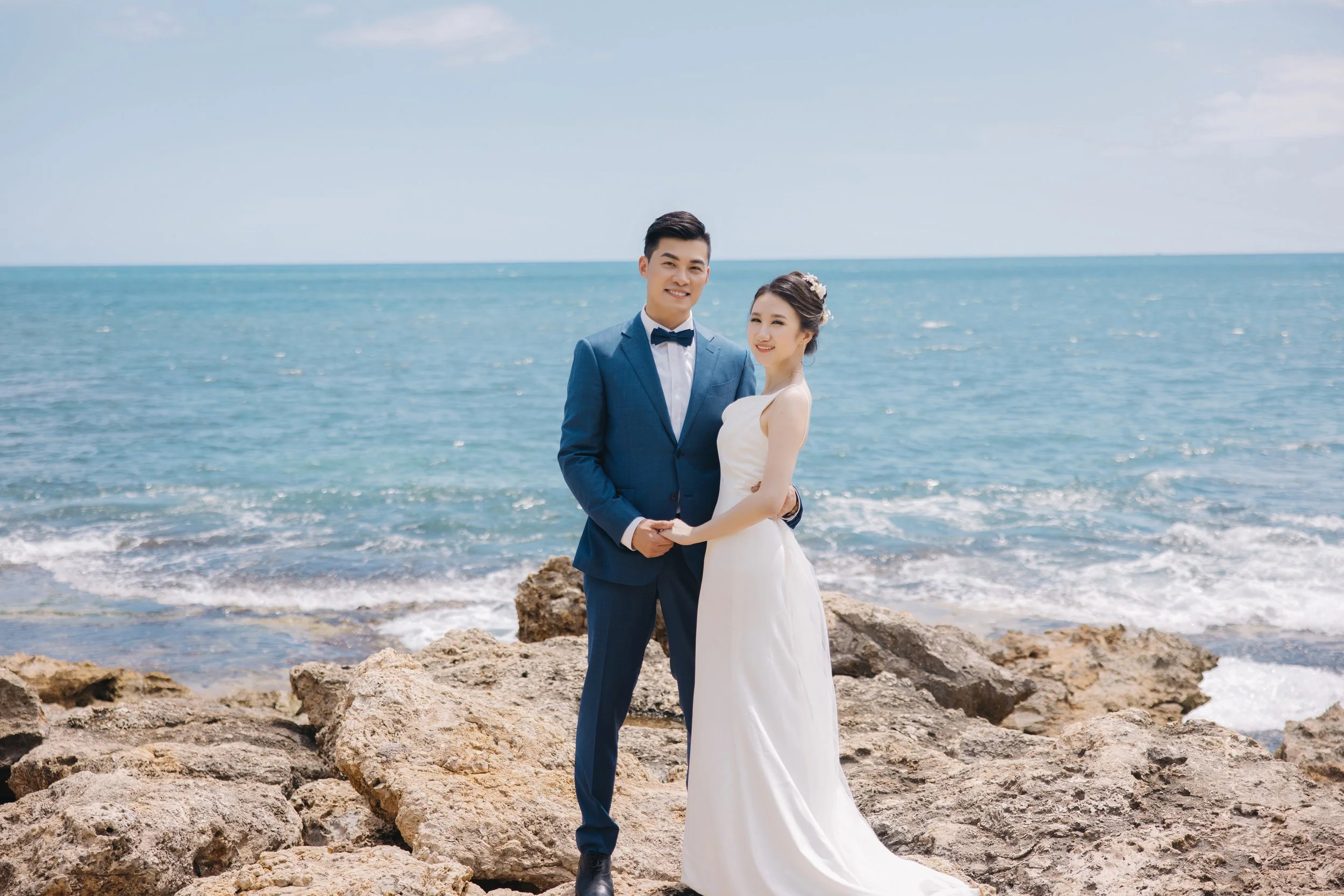 Couple in wedding attire standing on rocks by the ocean, holding hands, smiling, with a clear sky.