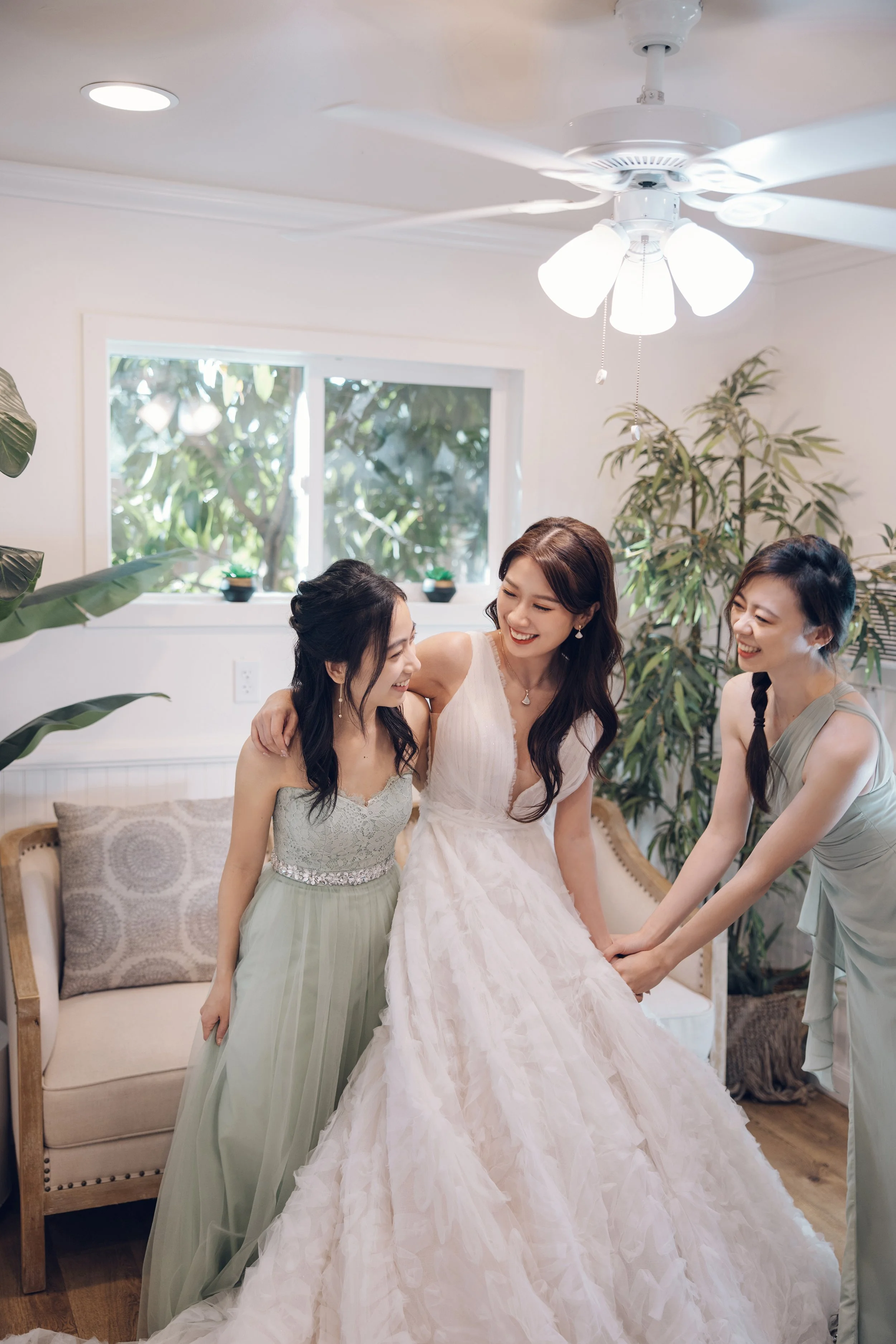 Three women, one in a white wedding dress and two in pastel dresses, smiling and helping the bride in a cozy room with green plants, a window, a ceiling fan, and a beige sofa.