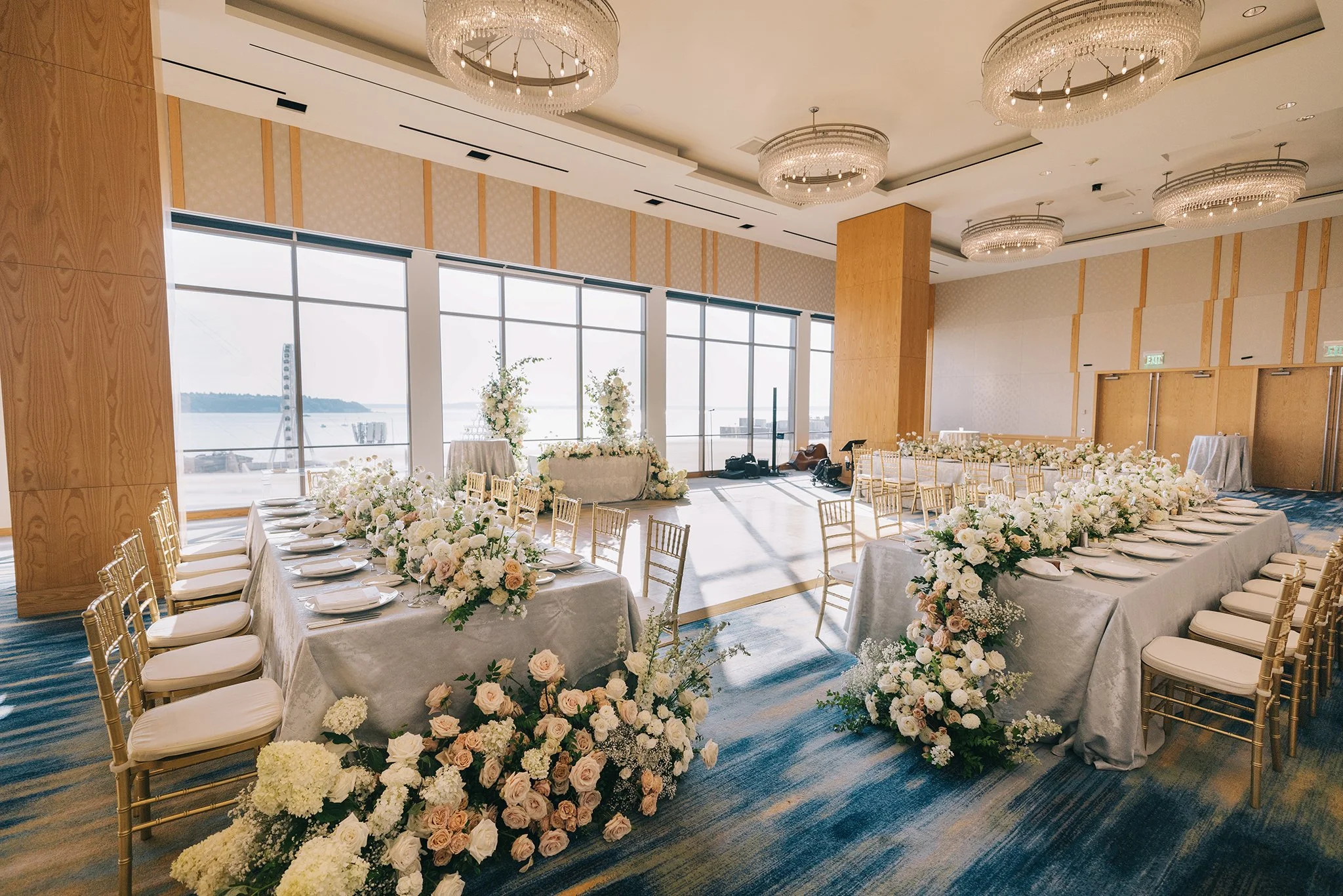Elegant wedding reception setup with long dining tables decorated with white flowers, gold chairs, and placed for guests. Large windows overlooking water in the background, chandeliers hanging from the ceiling, and floral arrangements along the table