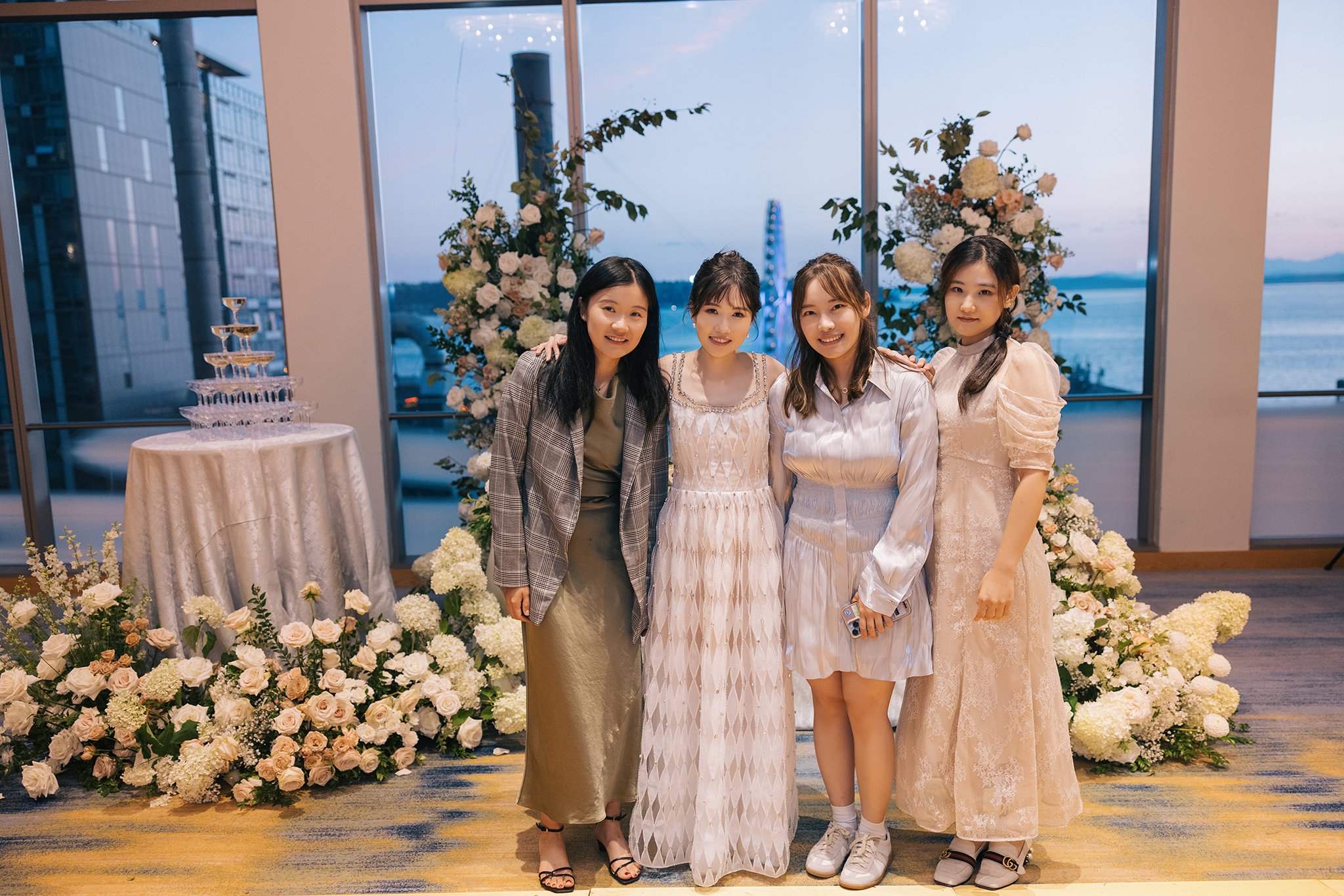 Four women standing together at an event with floral arrangements and a view of water and a ferris wheel in the background.