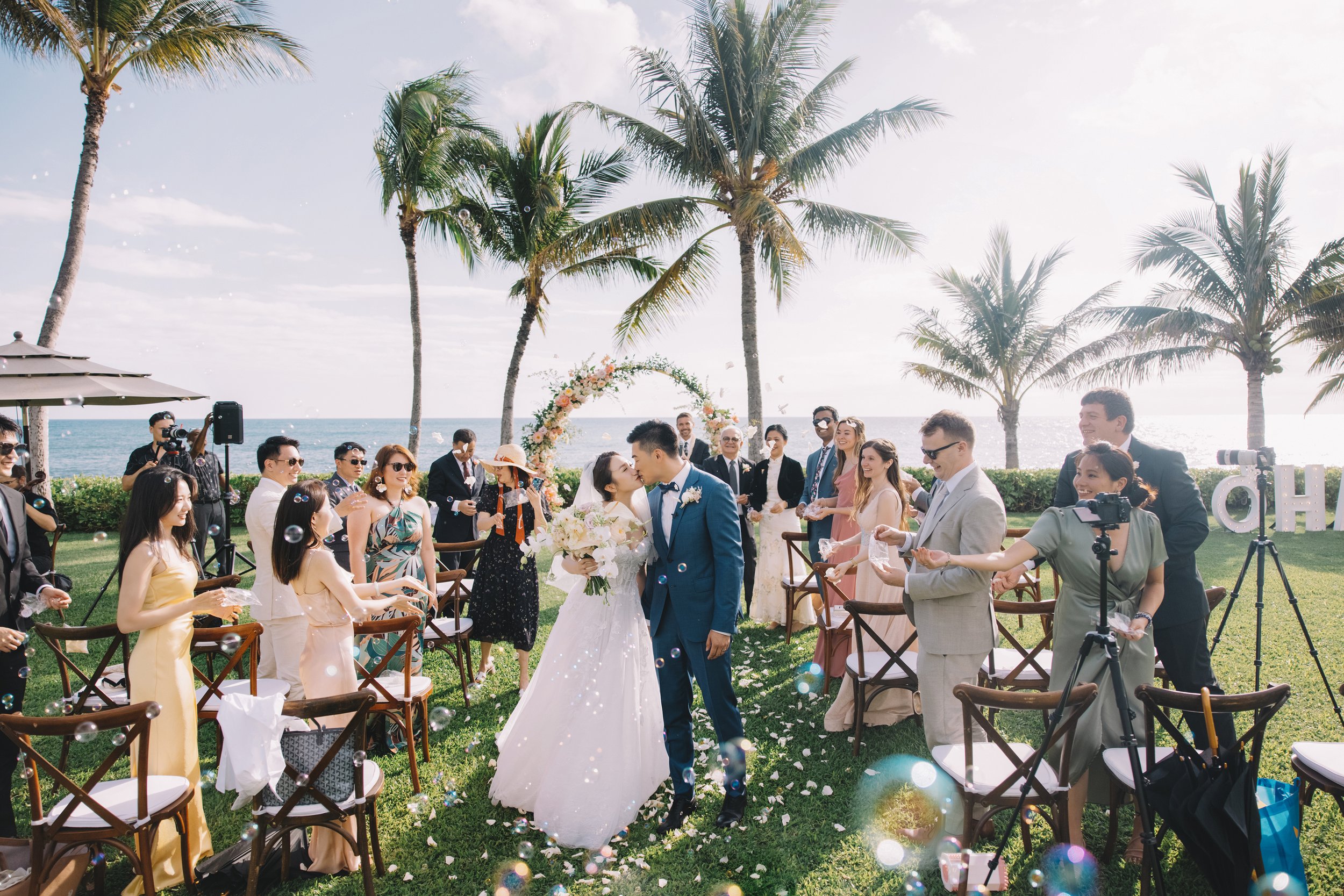 A wedding ceremony outdoors by the beach with the bride and groom kissing. Wedding guests are celebrating and throwing bubbles, with palm trees, the ocean, and a floral arch in the background.