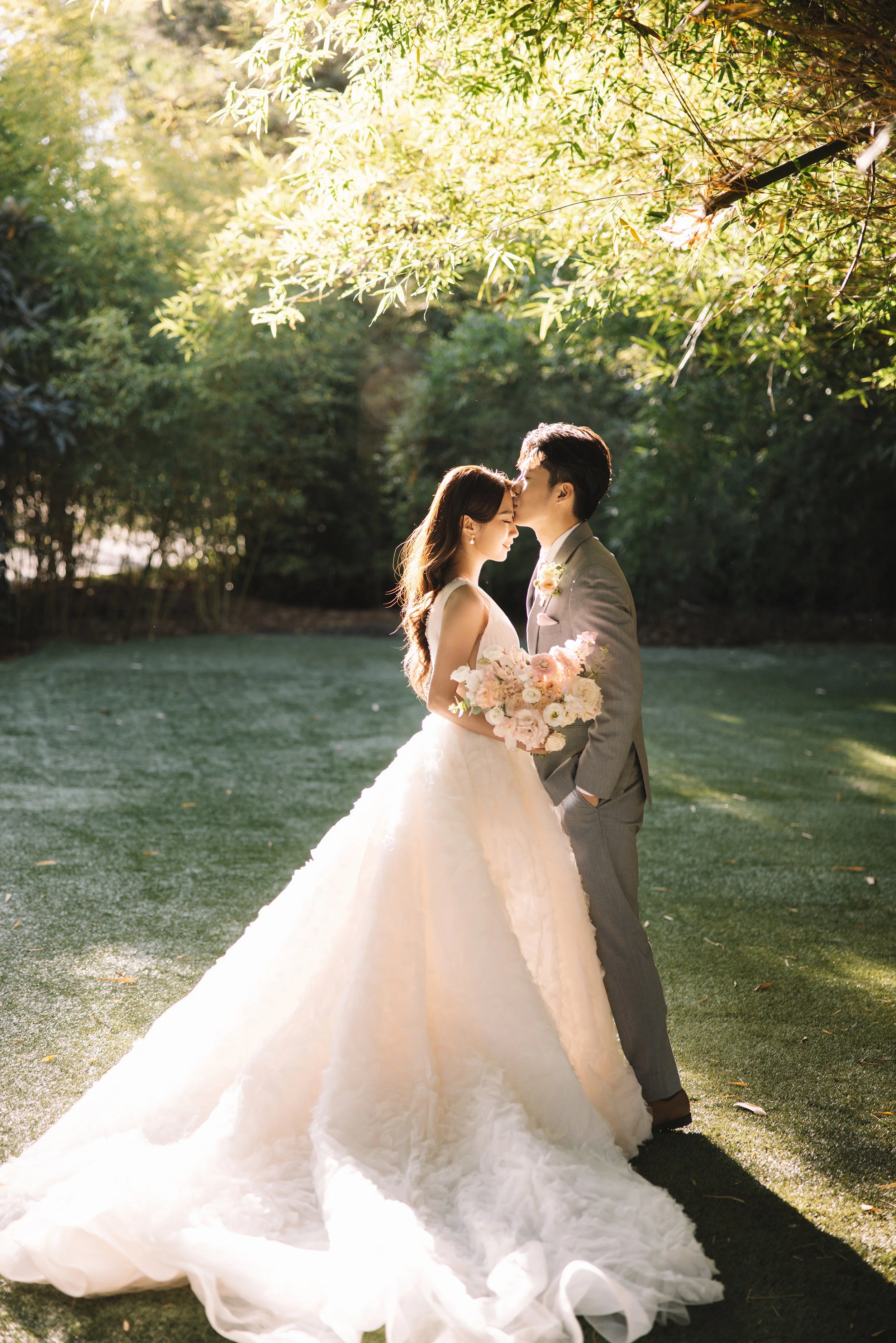 A bride and groom share a romantic kiss outdoors on a lush green lawn, with sunlight filtering through trees, the bride holding a bouquet of pink and white flowers.