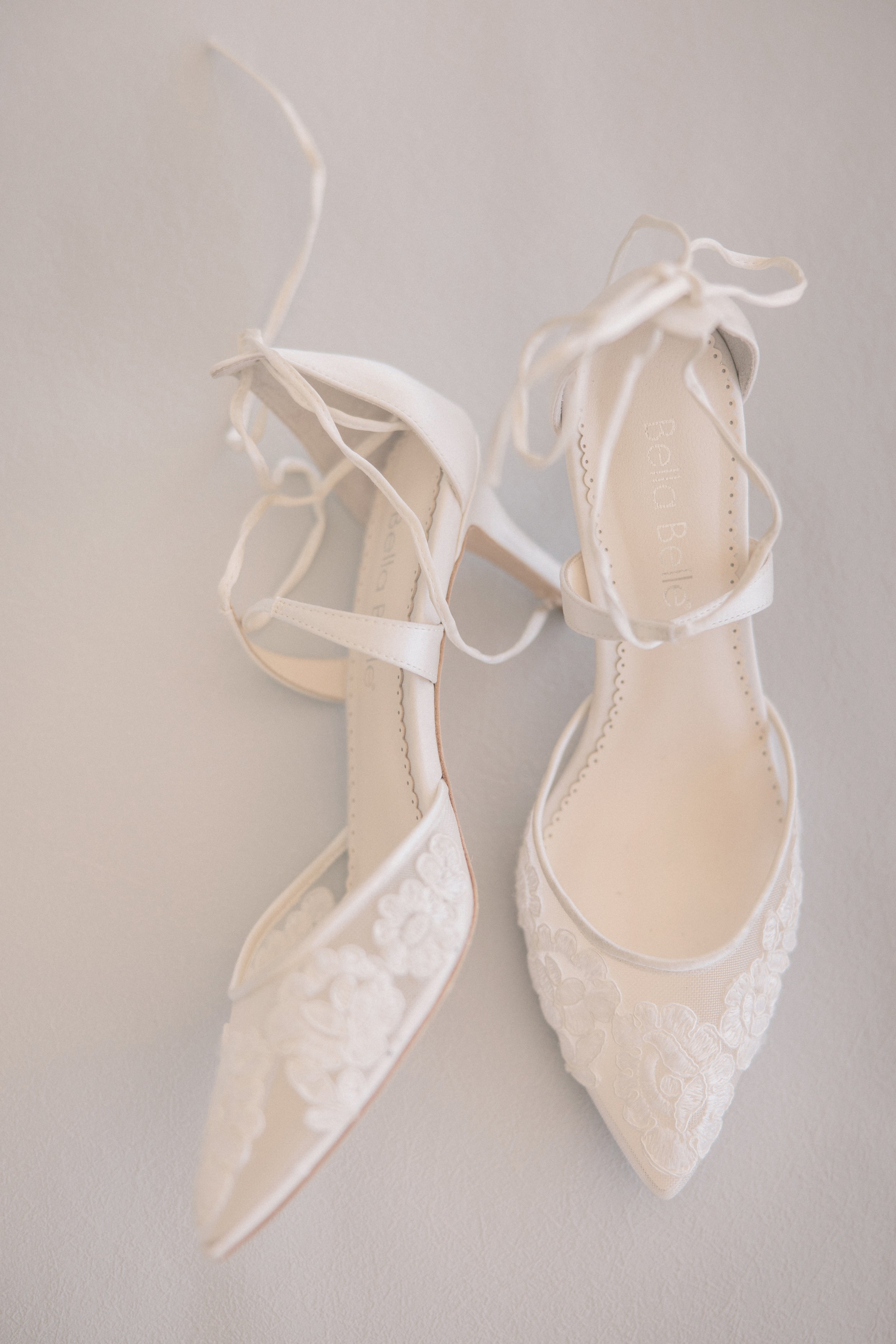 A pair of white lace wedding shoes with ankle straps hanging on a wall.