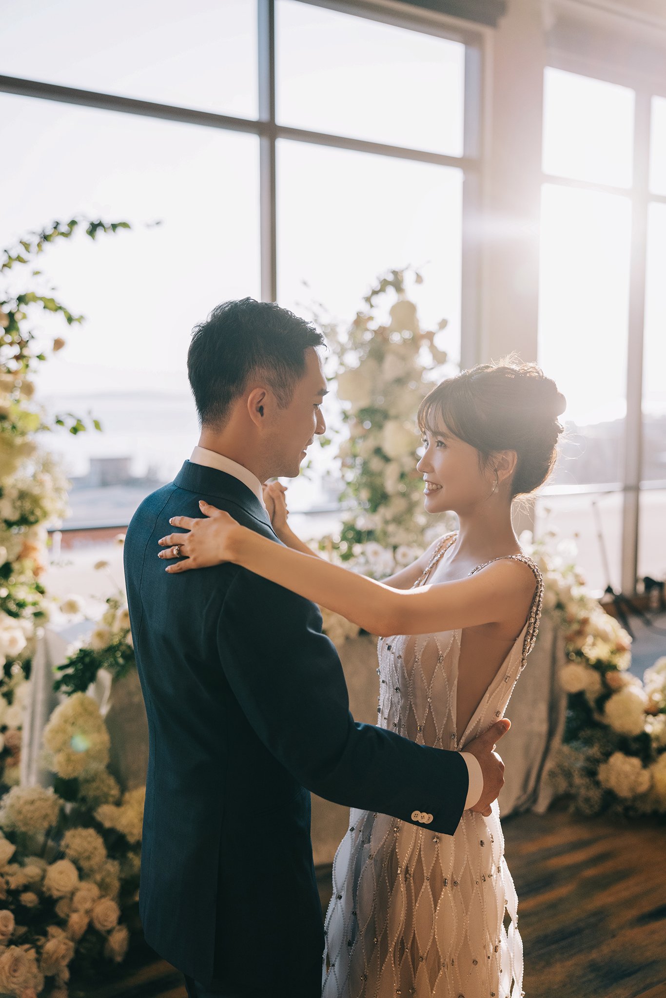 A bride and groom dancing indoors near a large window with sunlight, floral arrangements in the background.