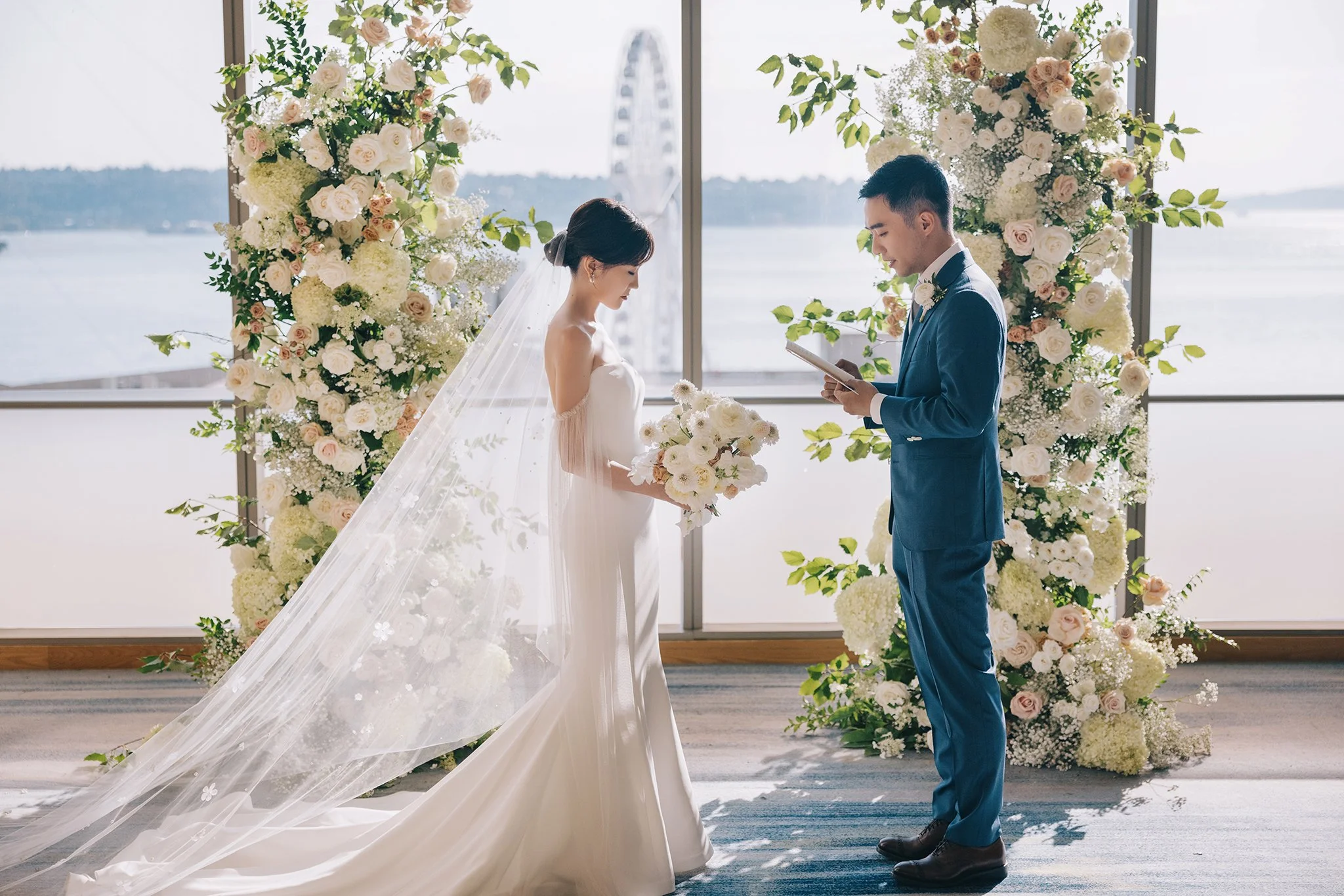 A bride and groom standing in front of wedding floral arch, exchanging vows during a wedding ceremony by a large window with a view of water and a Ferris wheel outside.