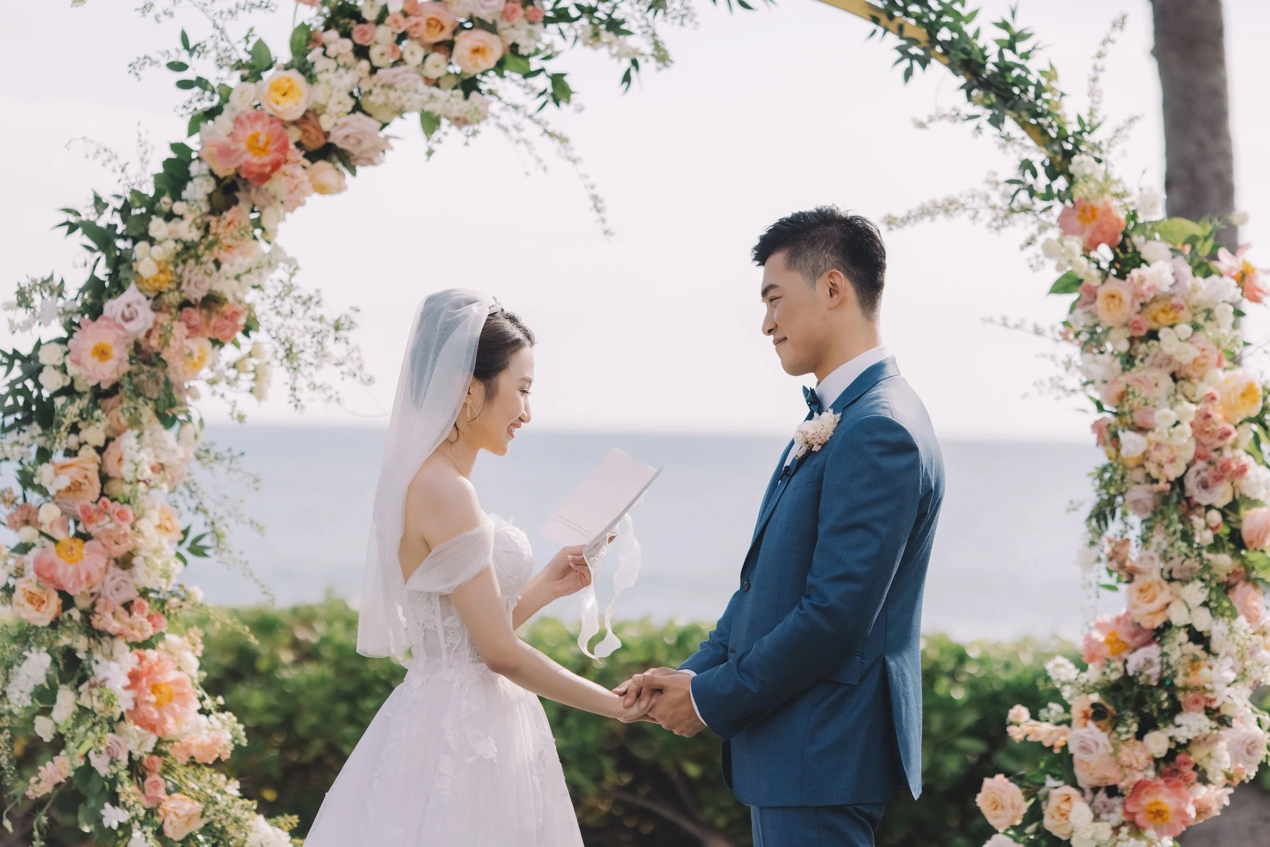 A bride and groom standing under a floral arch on a beach, holding hands and smiling at each other during their wedding ceremony.
