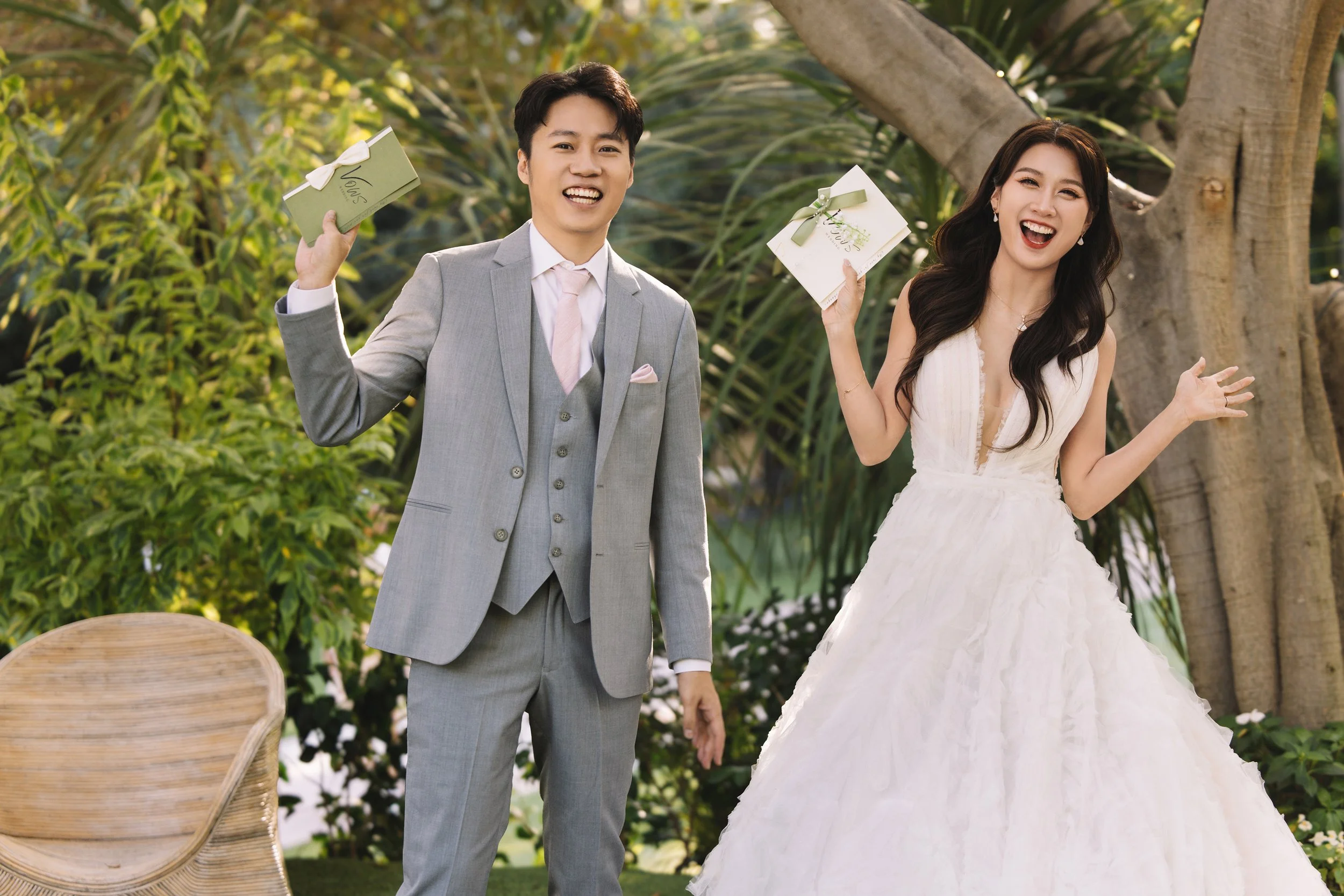 A happy couple at a wedding, holding gift boxes, outdoors with greenery and a tree in the background, the man in a gray suit and the woman in a white wedding dress.