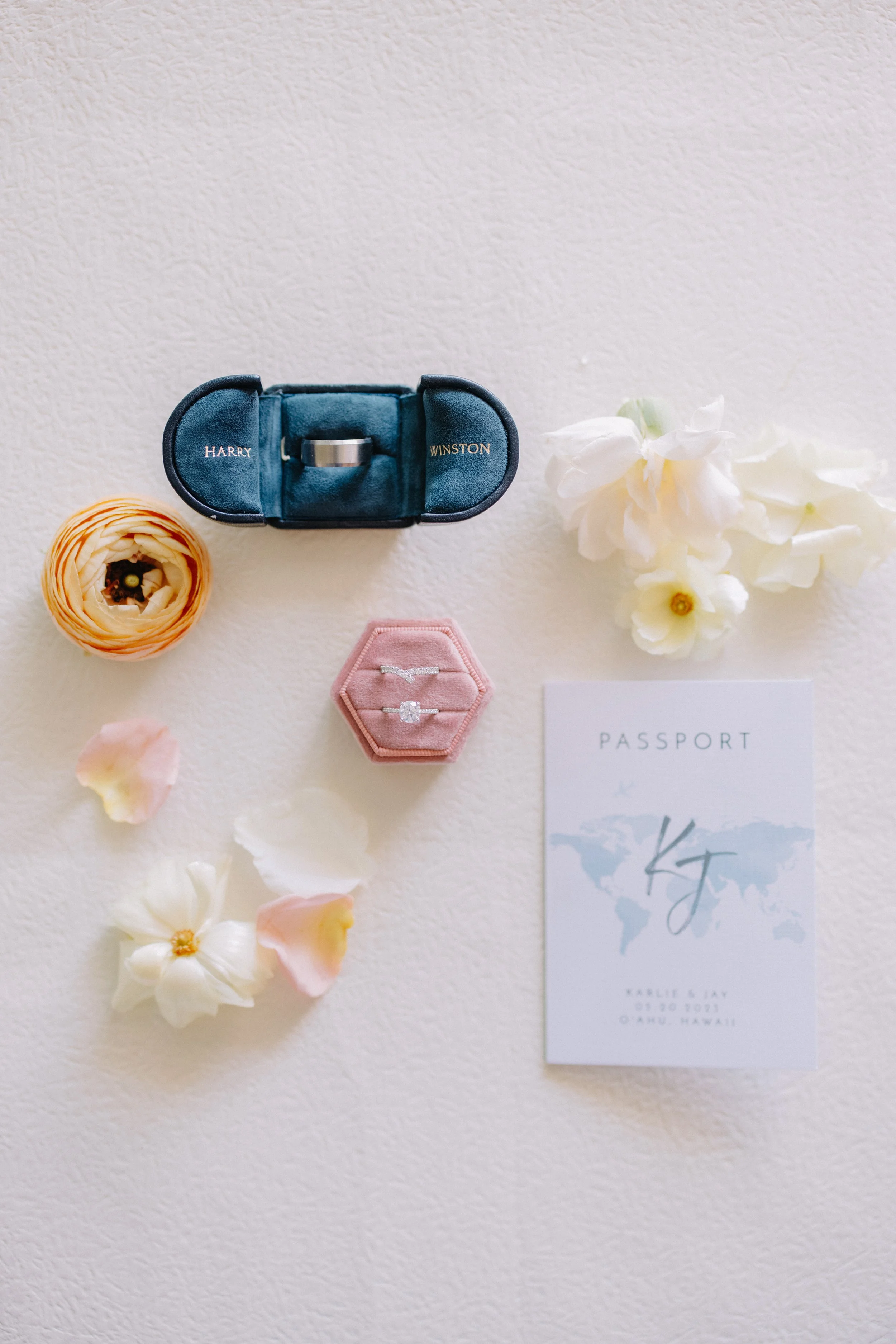 Wedding invitation with engagement ring and wedding bands, surrounded by flowers and petals, on a white surface.
