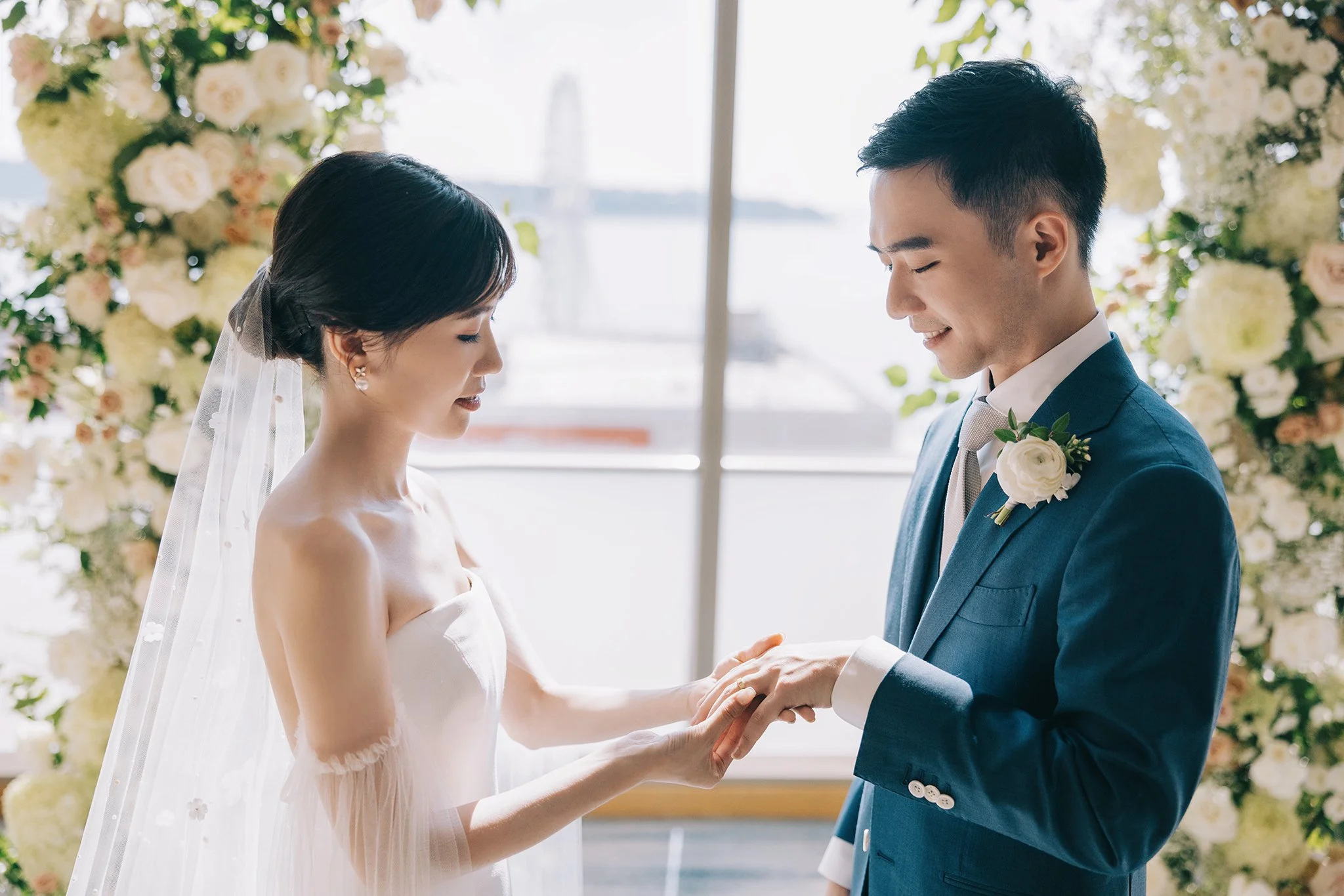 A bride and groom exchange rings during their wedding ceremony, standing in front of floral arrangements and with a window view of a bridge in the background.