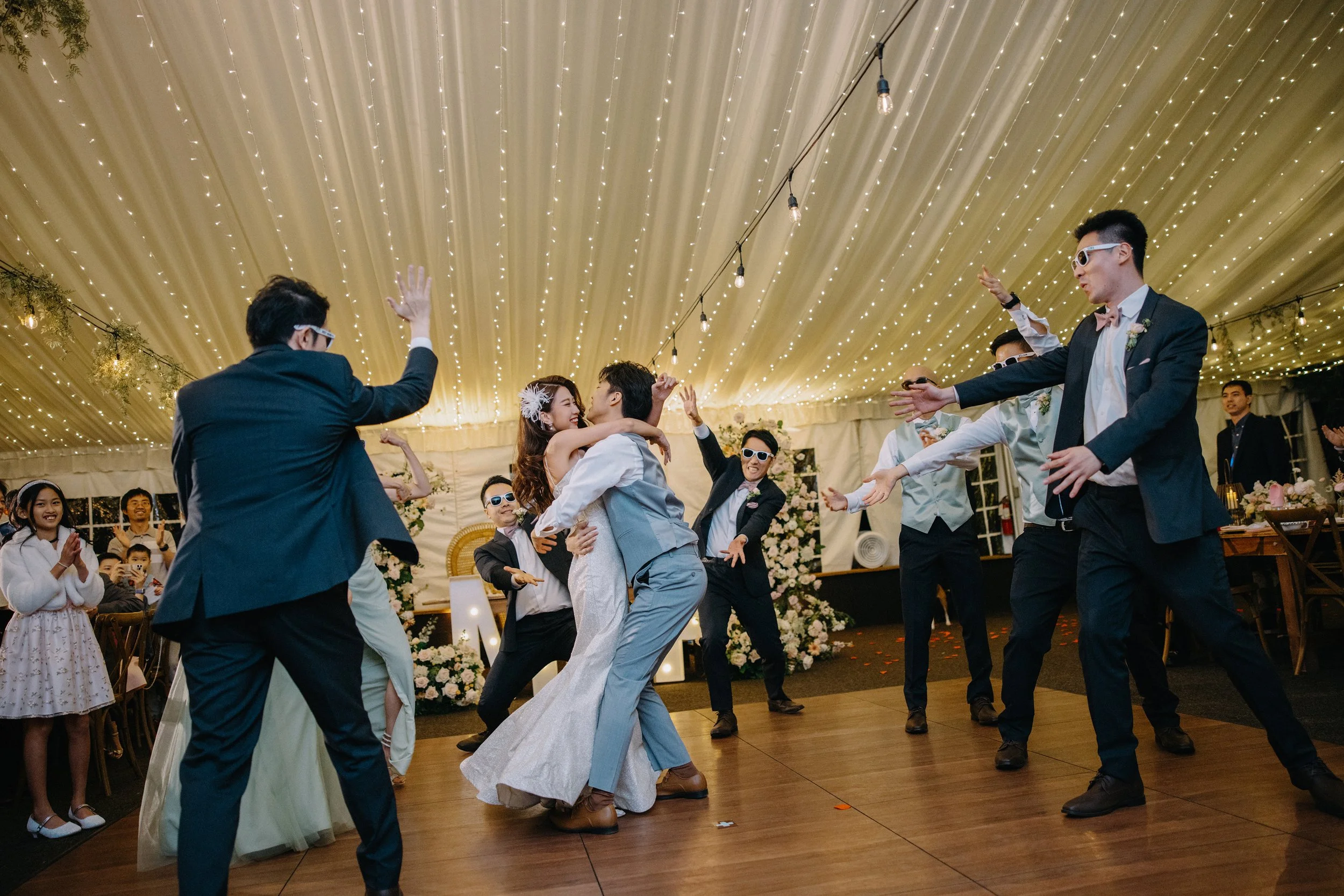 People dancing and celebrating at a wedding reception under a decorated tent with string lights and floral arrangements.