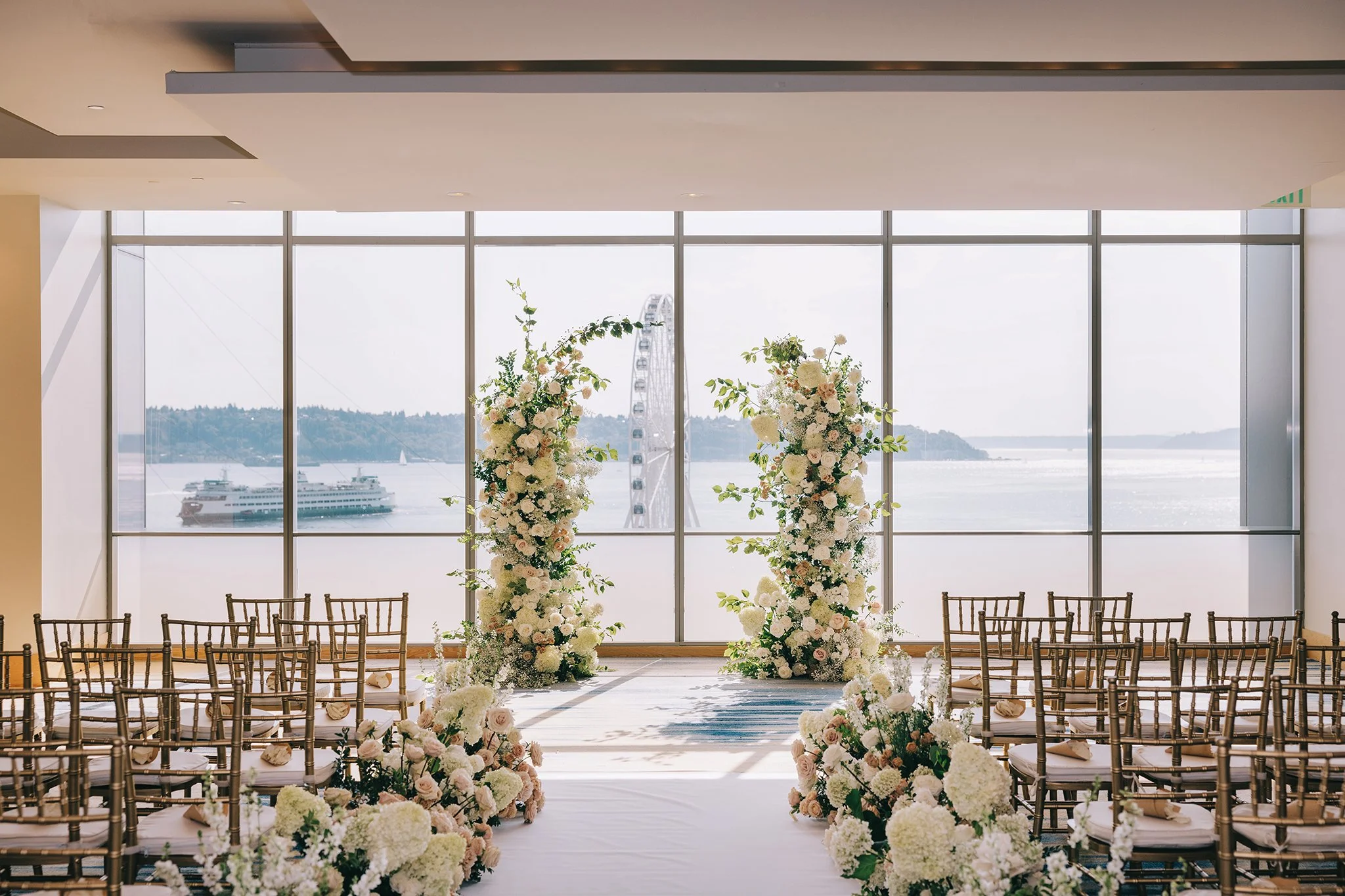 Indoor wedding ceremony setup with floral arch, chairs on either side, and a large window overlooking a body of water with boats and a Ferris wheel in the background.