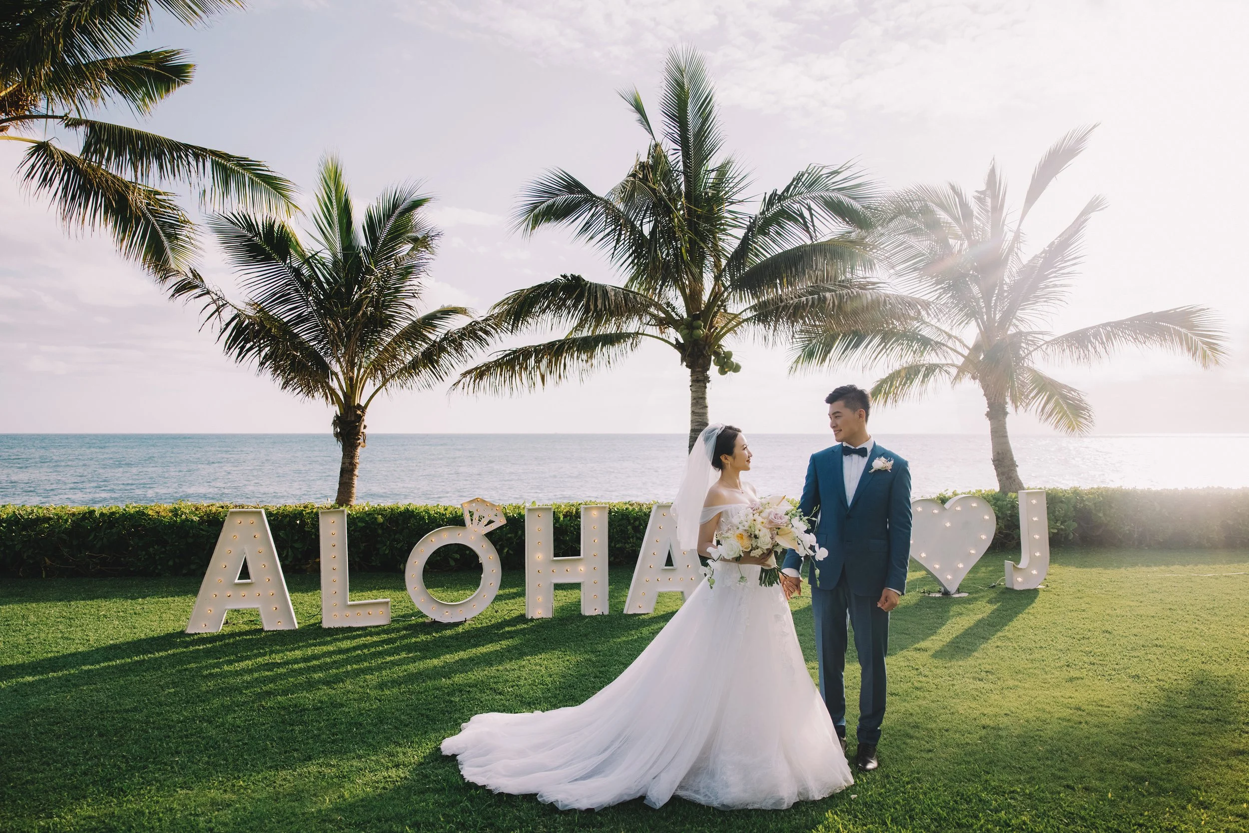 A bride and groom in wedding attire standing on a grassy area near the beach, holding hands and smiling at each other. Behind them are large decorative letters spelling "ALOHA" with a heart symbol, set against a backdrop of palm trees, ocean, and a p