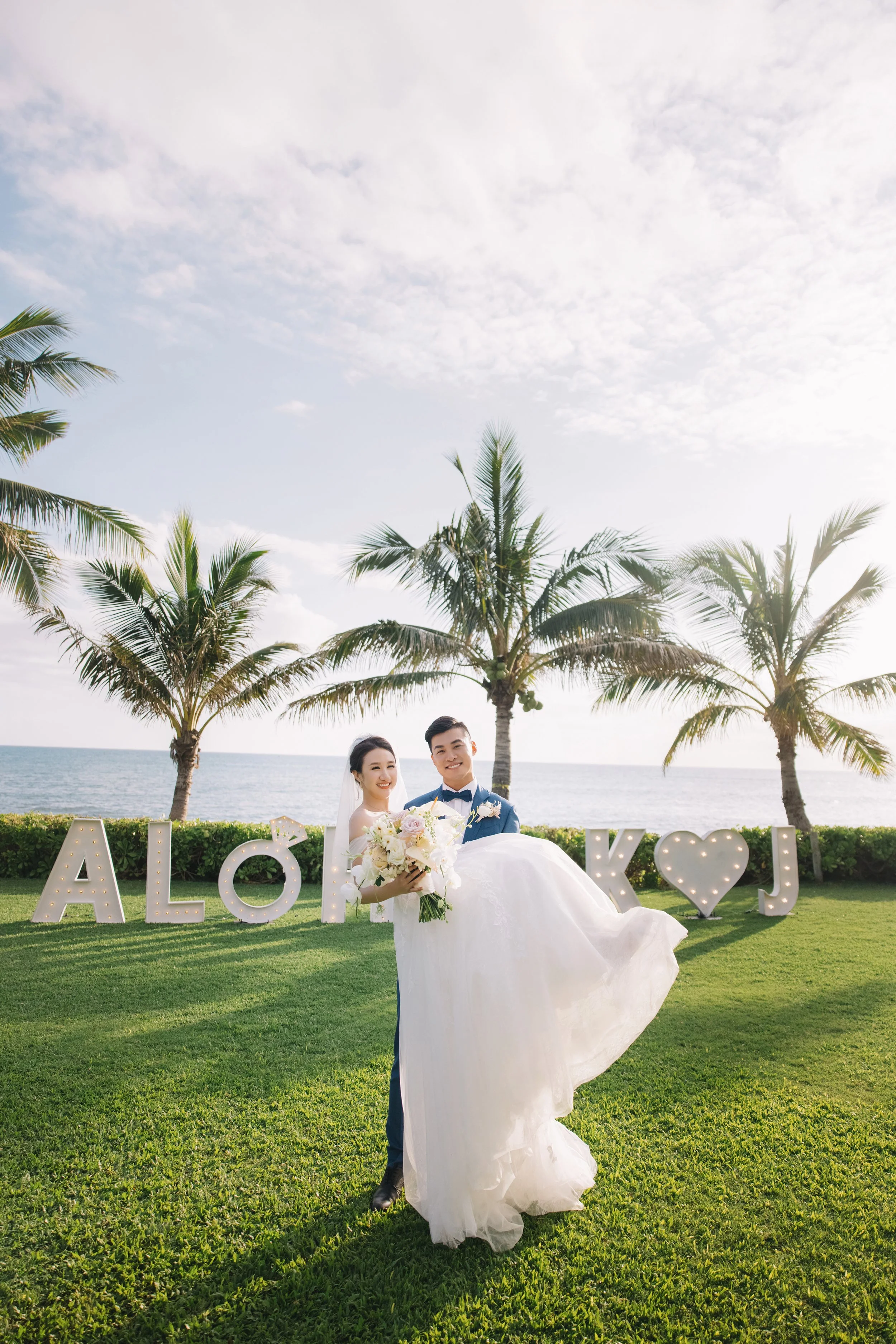 A newlywed couple outdoors with palm trees in the background, the groom carrying the bride as they smile for the camera in front of illuminated signs saying 'ALOHA' and a heart symbol.
