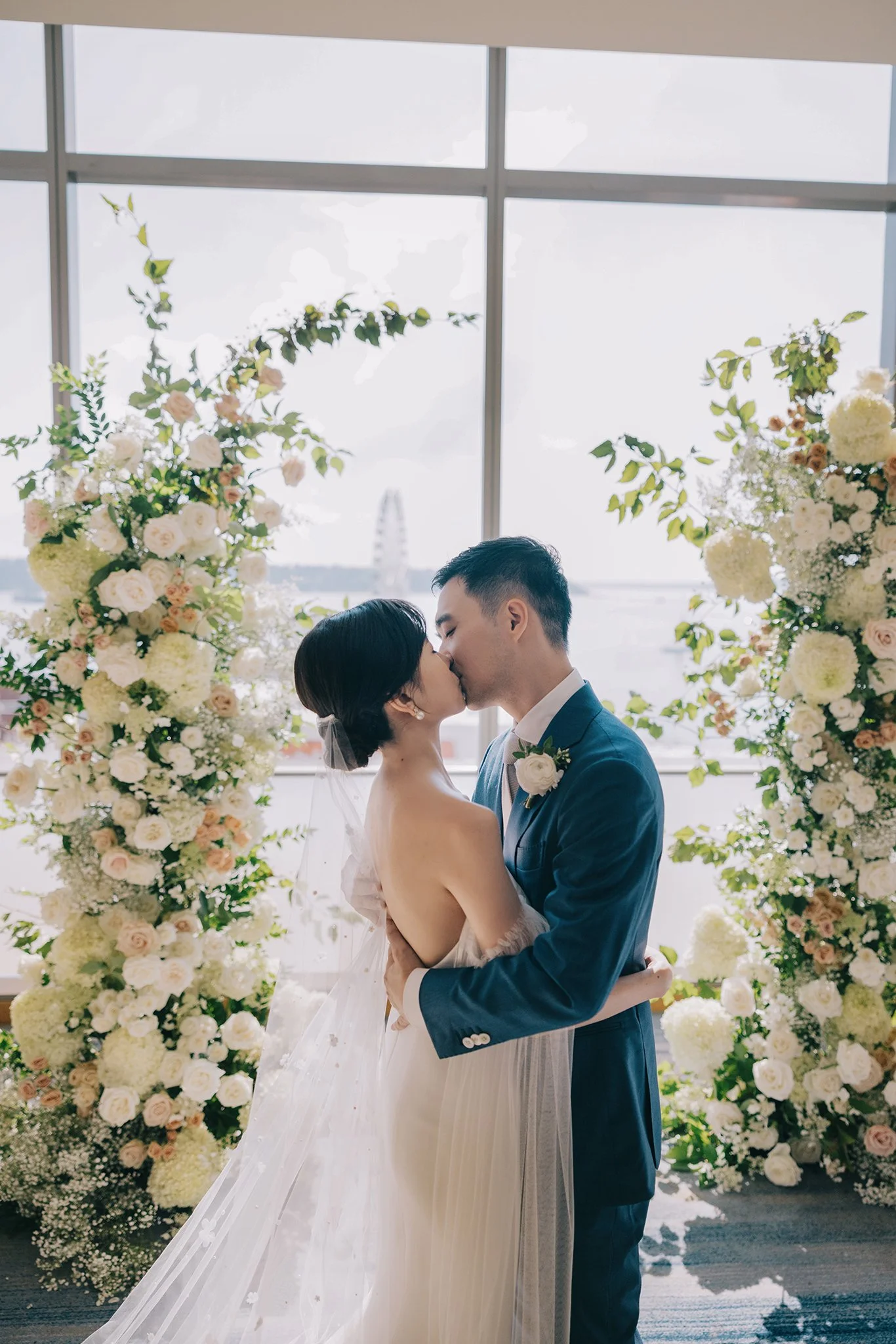 A bride and groom share a kiss during their wedding ceremony, standing between large floral arrangements, with a window and water view in the background.