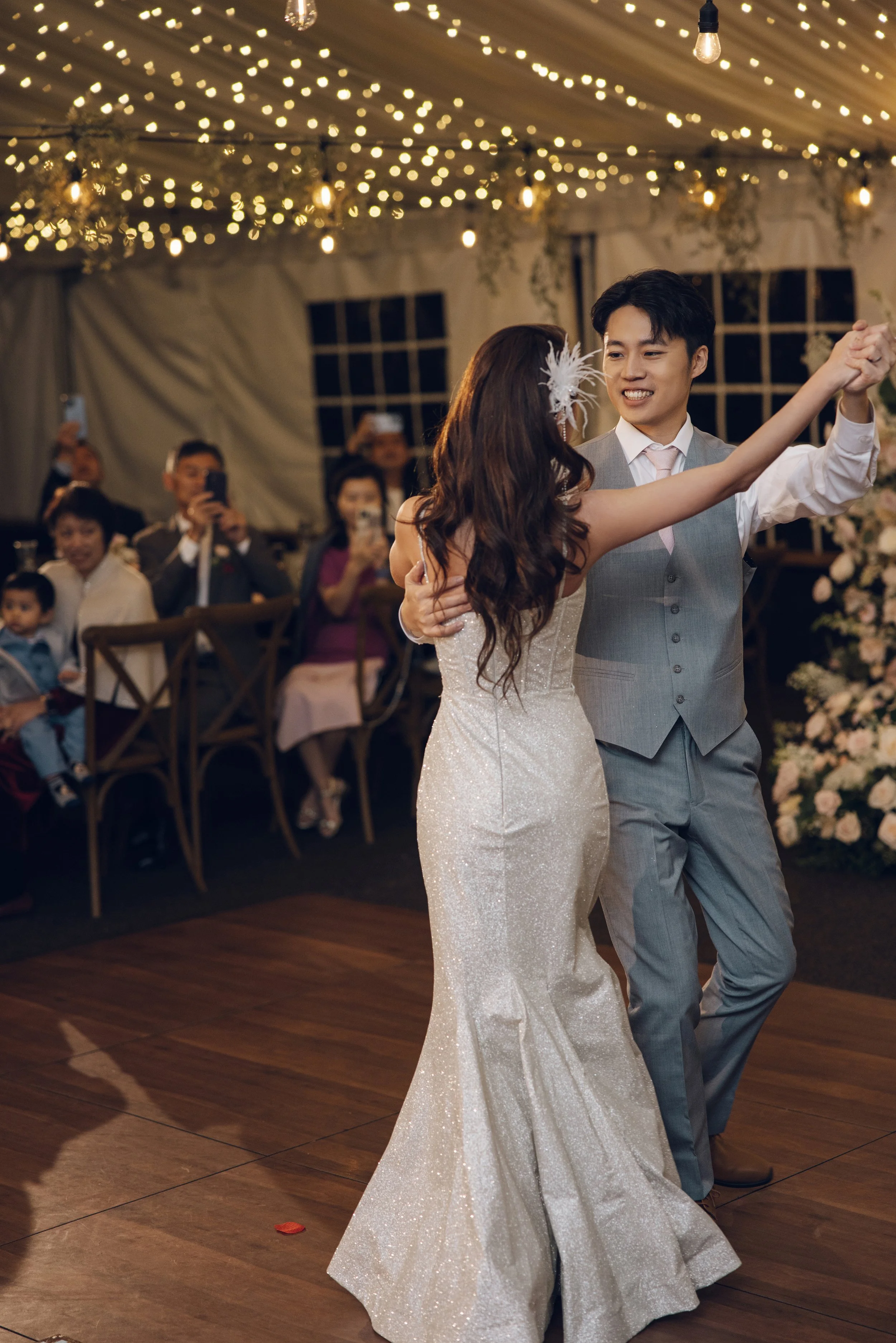 A bride and groom are dancing at their wedding reception under string lights and surrounded by guests, some of whom are taking photos.