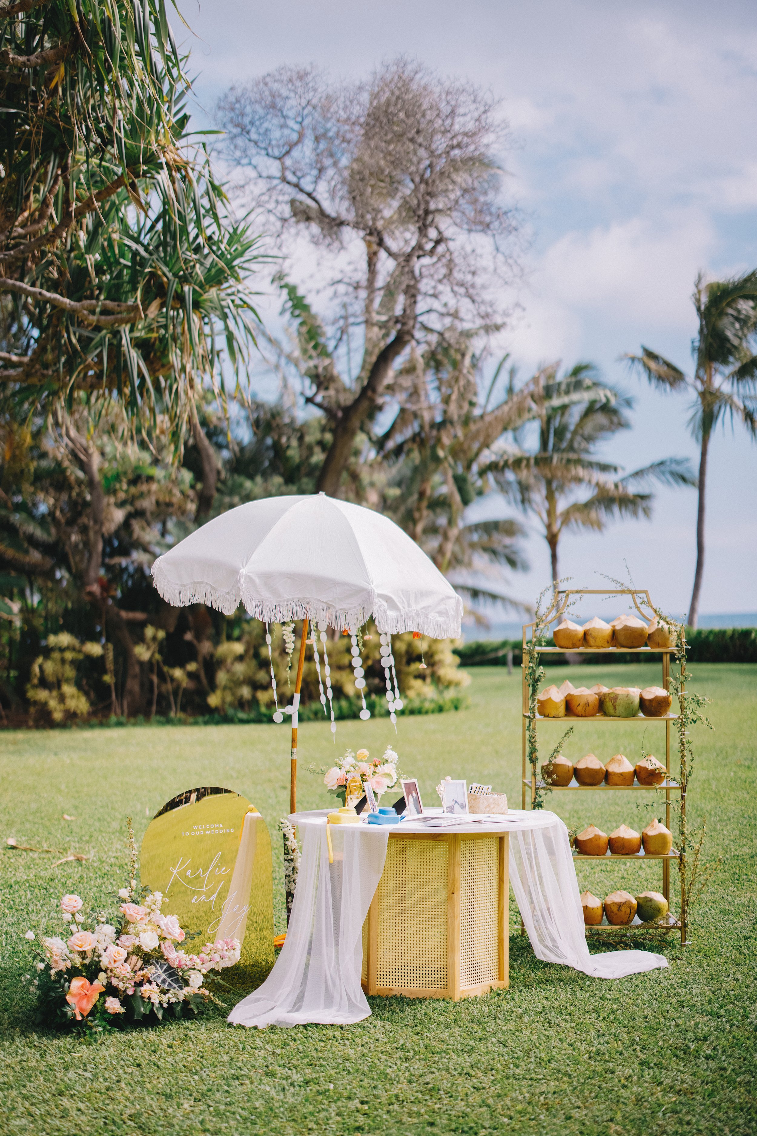 Decorative outdoor scene with a white parasol decorated with hanging white beads, a table with a white cloth, framed photos, flowers, and small boxes, a gold shelving unit holding coconuts, and a floral wreath and wooden sign with names on the grassy