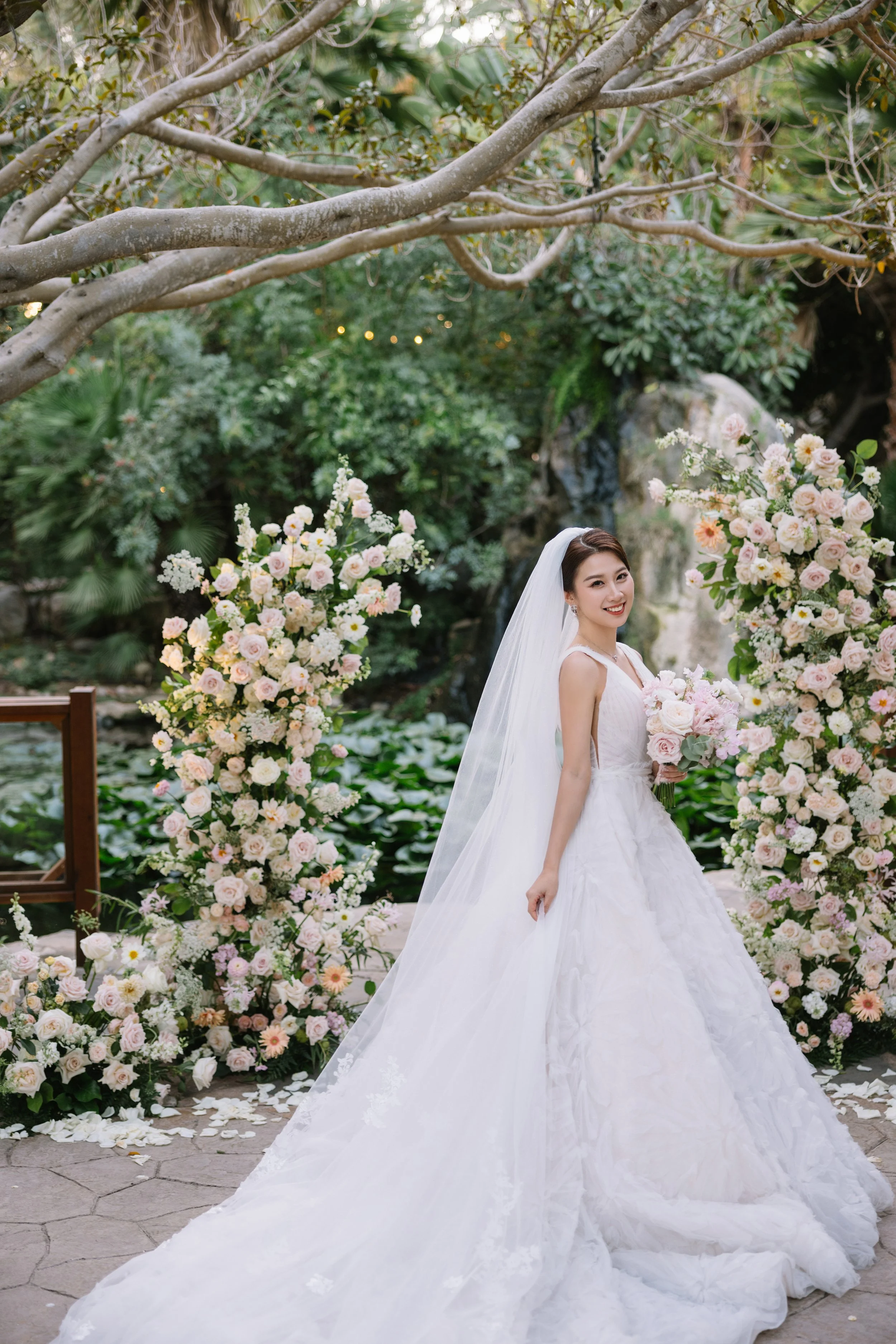 A smiling bride in a white wedding gown holding a bouquet of pink and white roses, standing outdoors in front of a floral arch and surrounded by lush greenery.