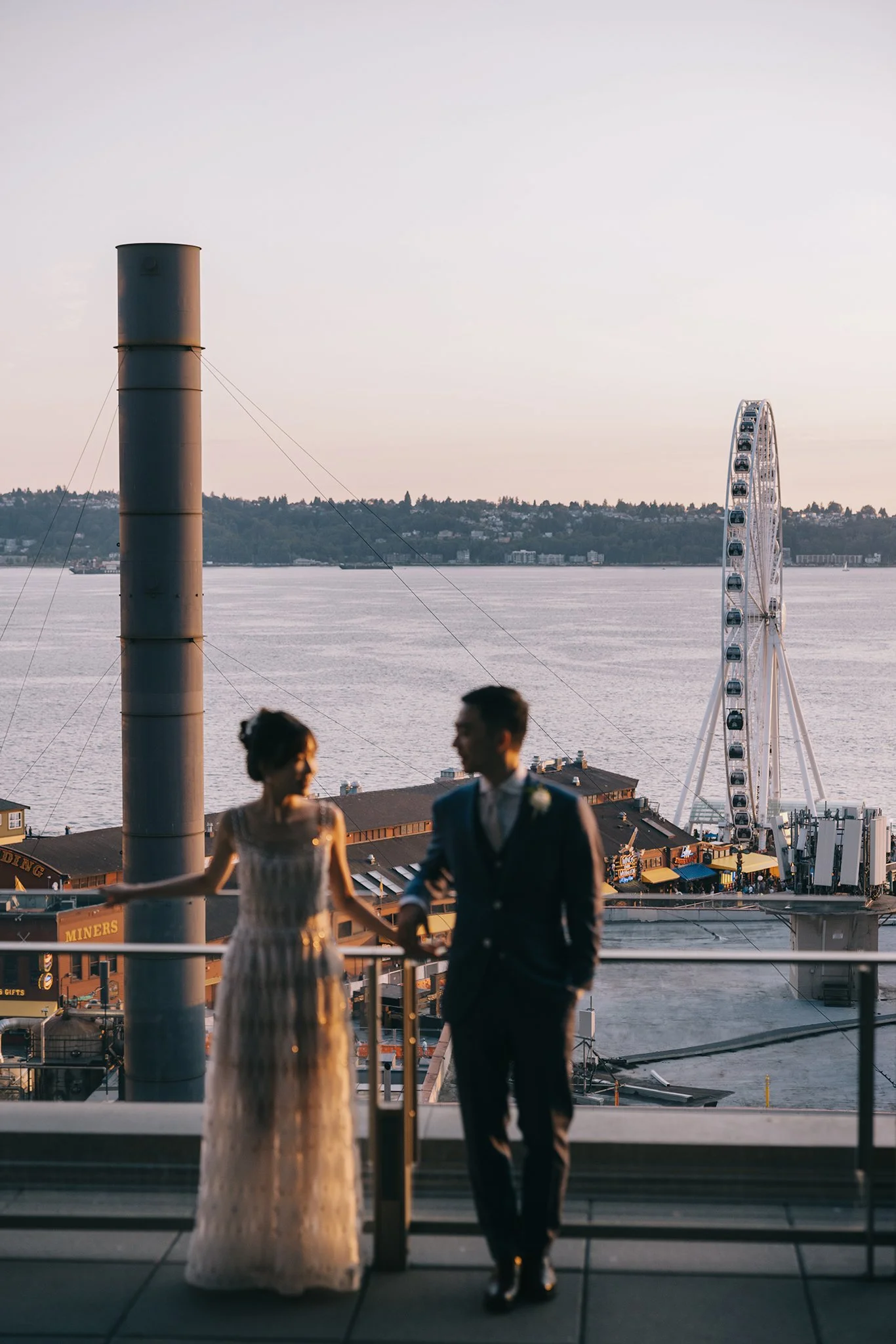A couple in wedding attire standing on a balcony with a ferris wheel and waterfront view in the background.