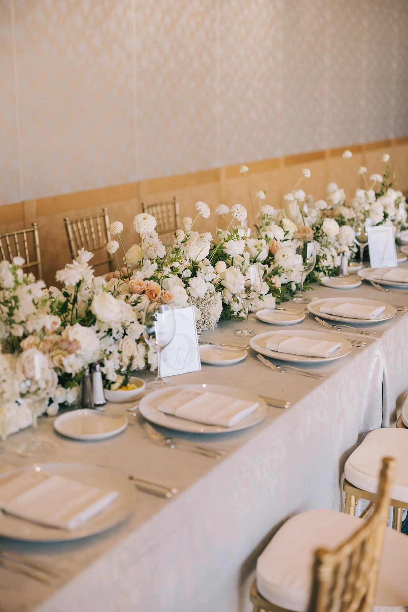 A long dining table decorated with white and blush pink flowers and tableware, set for a formal event or wedding reception in a well-lit room.