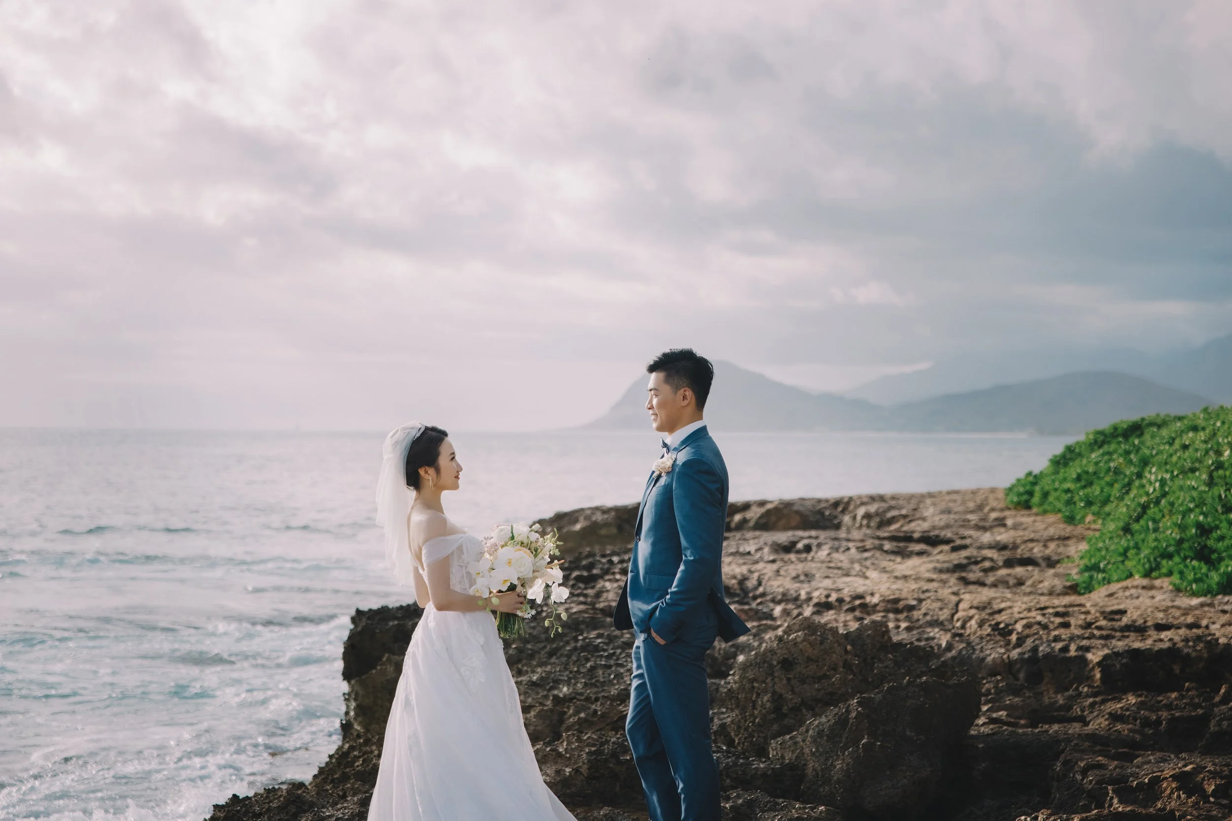 A bride and groom standing on a rocky shore by the ocean, facing each other. The bride is holding a bouquet of white flowers and wearing a white wedding dress with an off-shoulder neckline and a veil. The groom is dressed in a blue suit with a bow ti