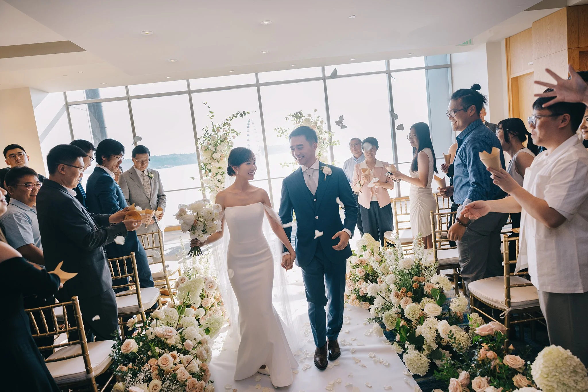 Bride and groom walk down the aisle hand-in-hand during their wedding ceremony, surrounded by seated guests and floral decorations, with large windows and a view outside.