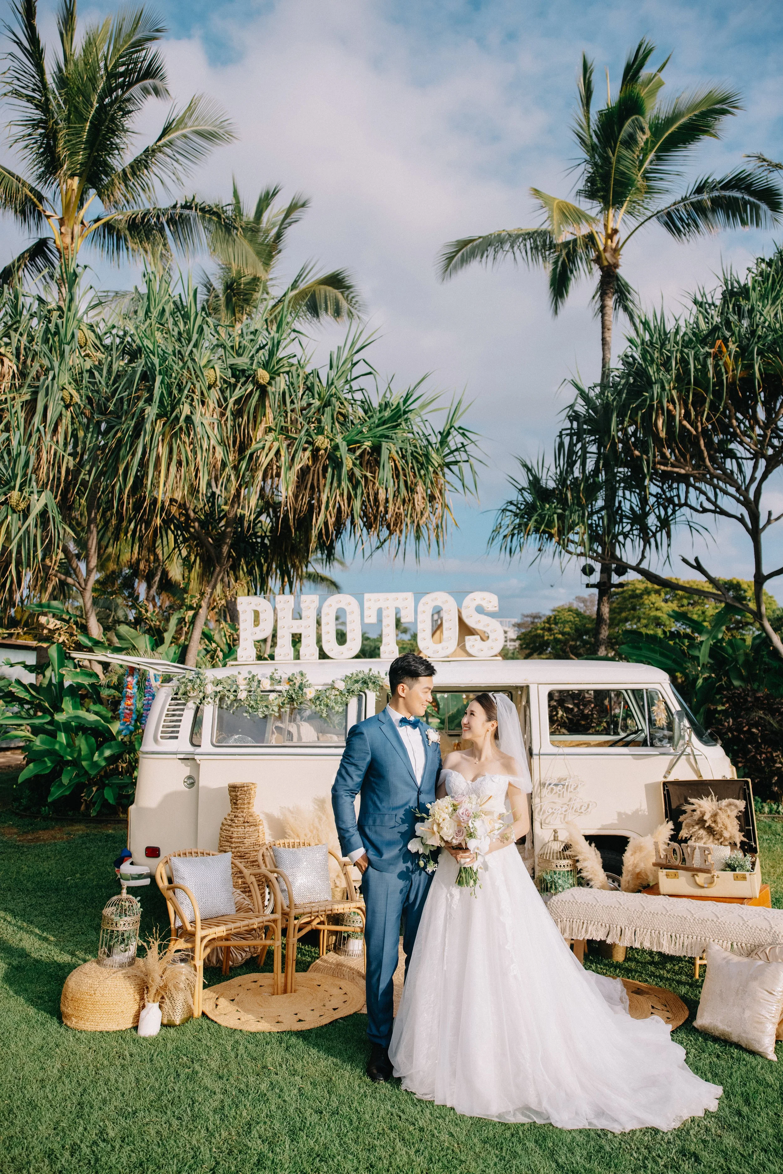 Bride and groom standing in front of a vintage van decorated for a wedding, with large illuminated 'PHOTOS' sign on top, surrounded by tropical greenery and outdoor decor.
