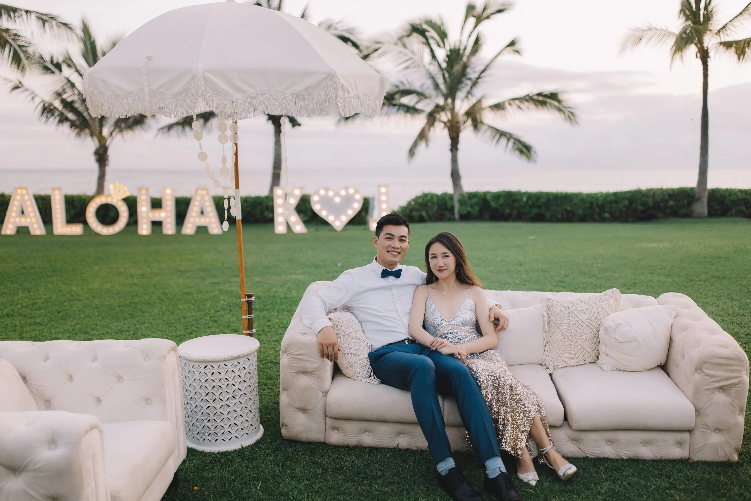 A couple sitting on a white sofa outdoors by the beach during sunset. The man is wearing a white shirt with a bow tie, and the woman is wearing a silver sequin dress. Behind them, there are large light-up letters spelling 'ALOHA' and 'K' with a heart