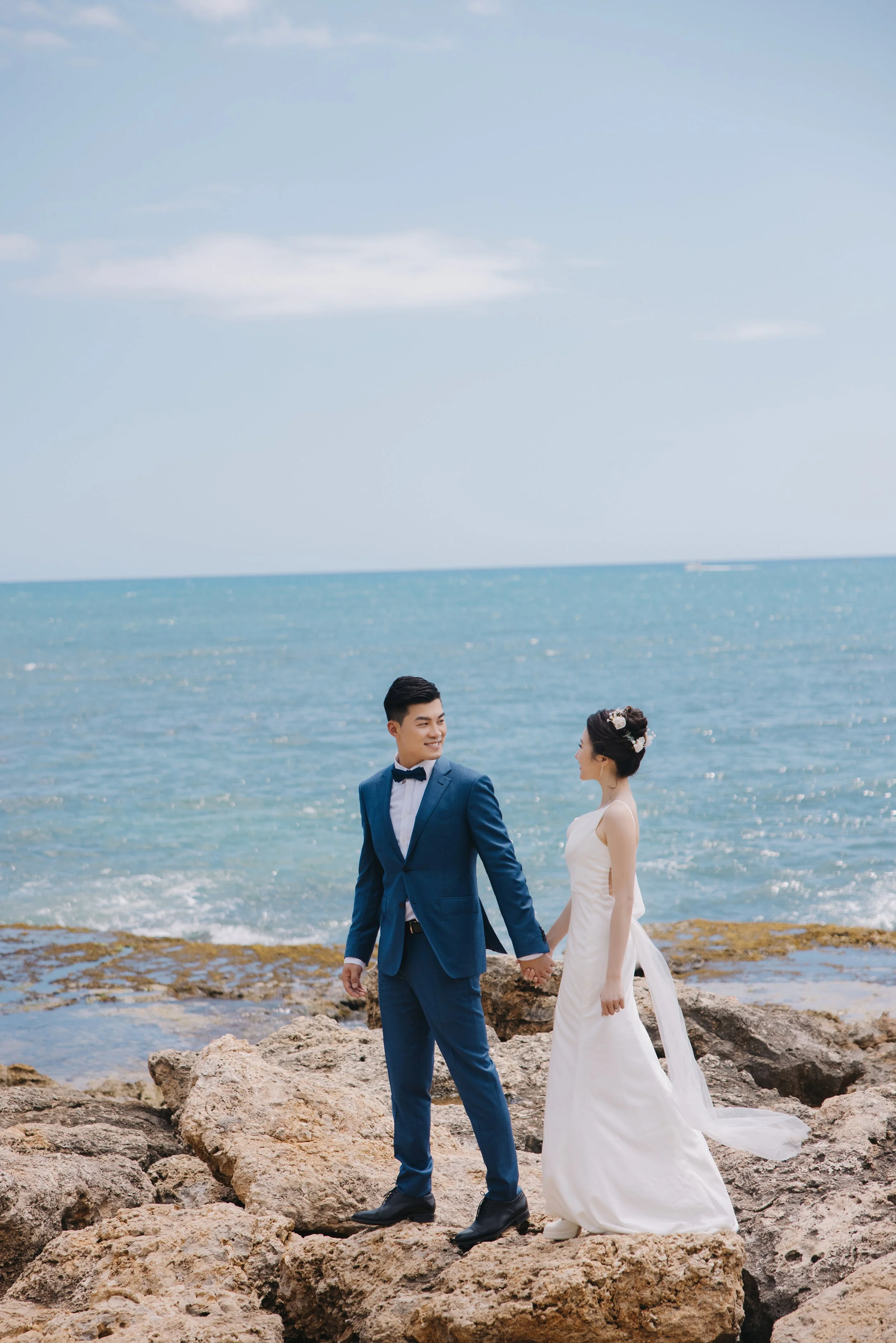 A bride and groom holding hands on rocks by the ocean on a sunny day, with the bride in a white wedding gown and the groom in a blue suit.