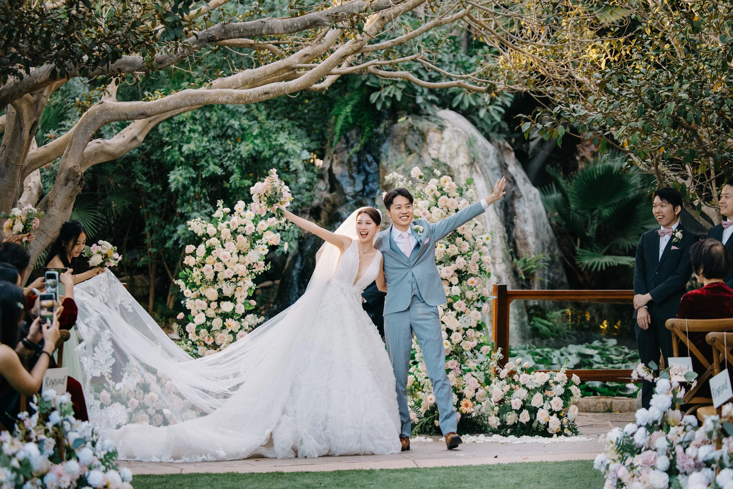 A newlywed couple celebrating their wedding outdoors with friends and family, surrounded by floral decorations and greenery.