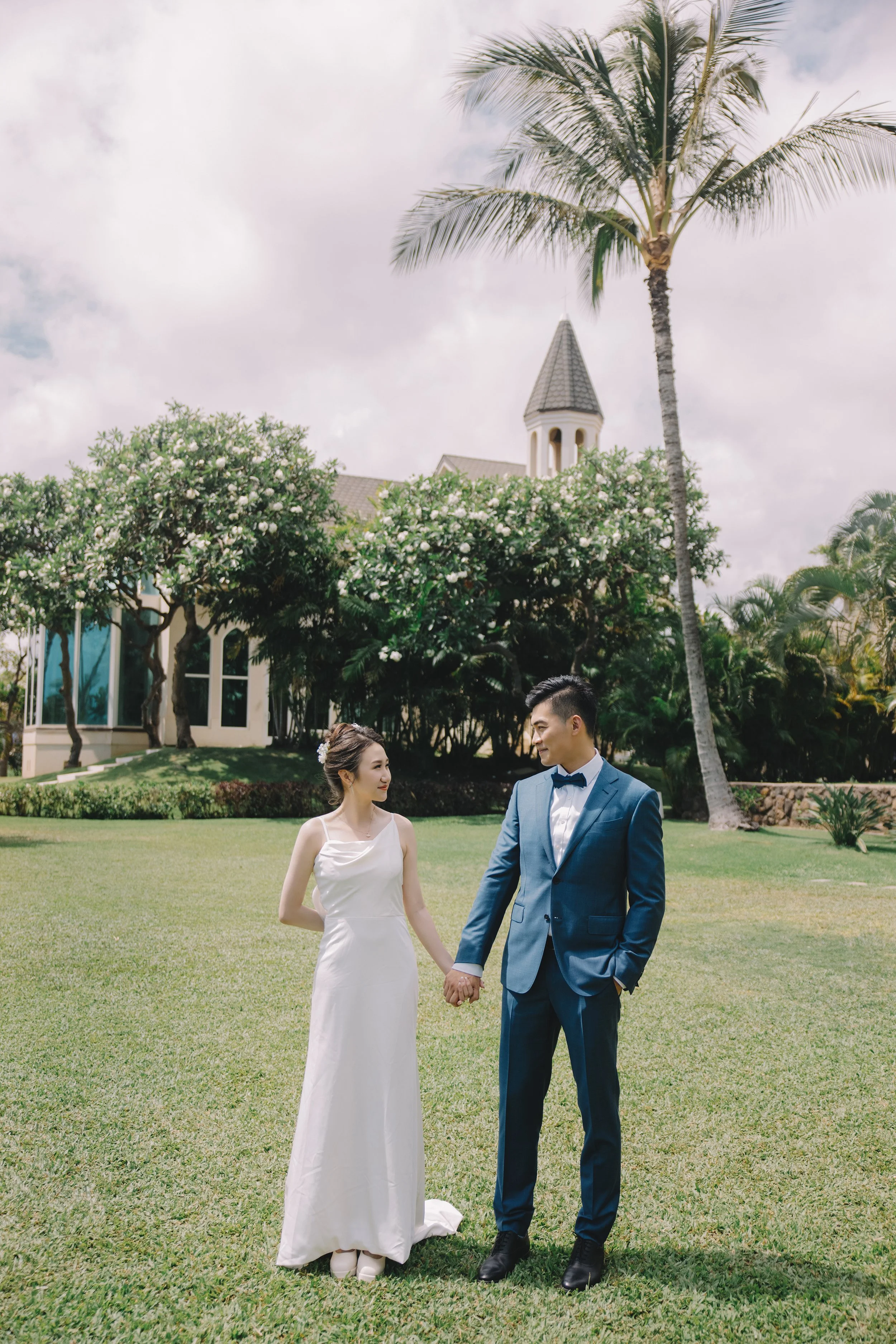 A bride and groom holding hands outdoors on grass, with a church, trees, and a palm tree in the background.