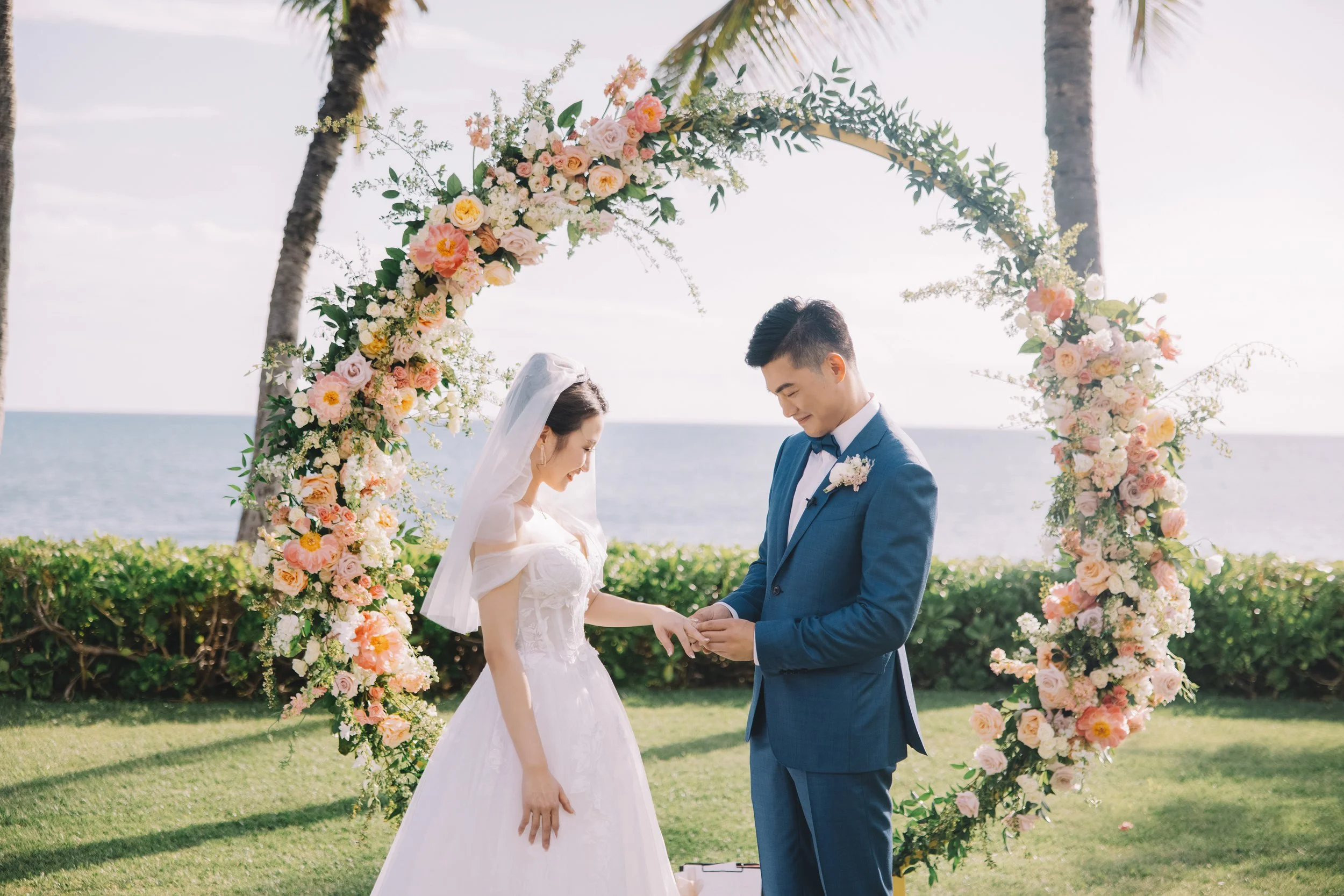 A bride and groom exchanging rings during an outdoor wedding ceremony by the ocean, with a floral arch and palm trees.