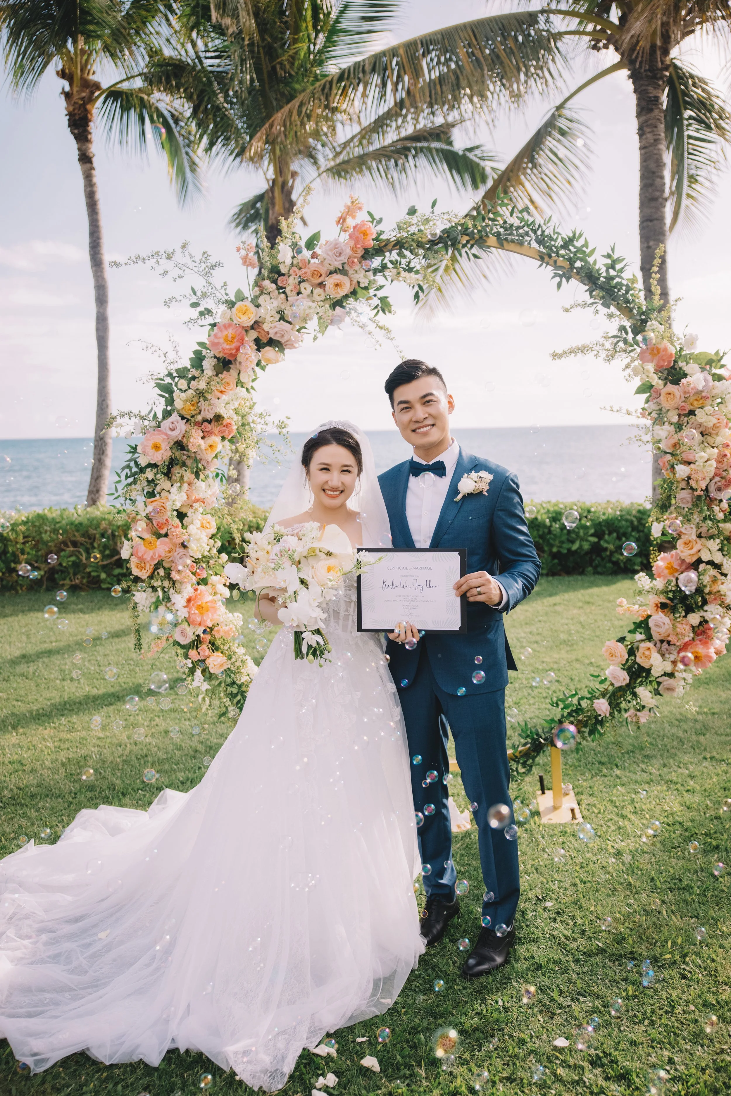 A newlywed couple in wedding attire standing under a floral arch on a grassy area near the ocean, smiling and holding a marriage certificate.
