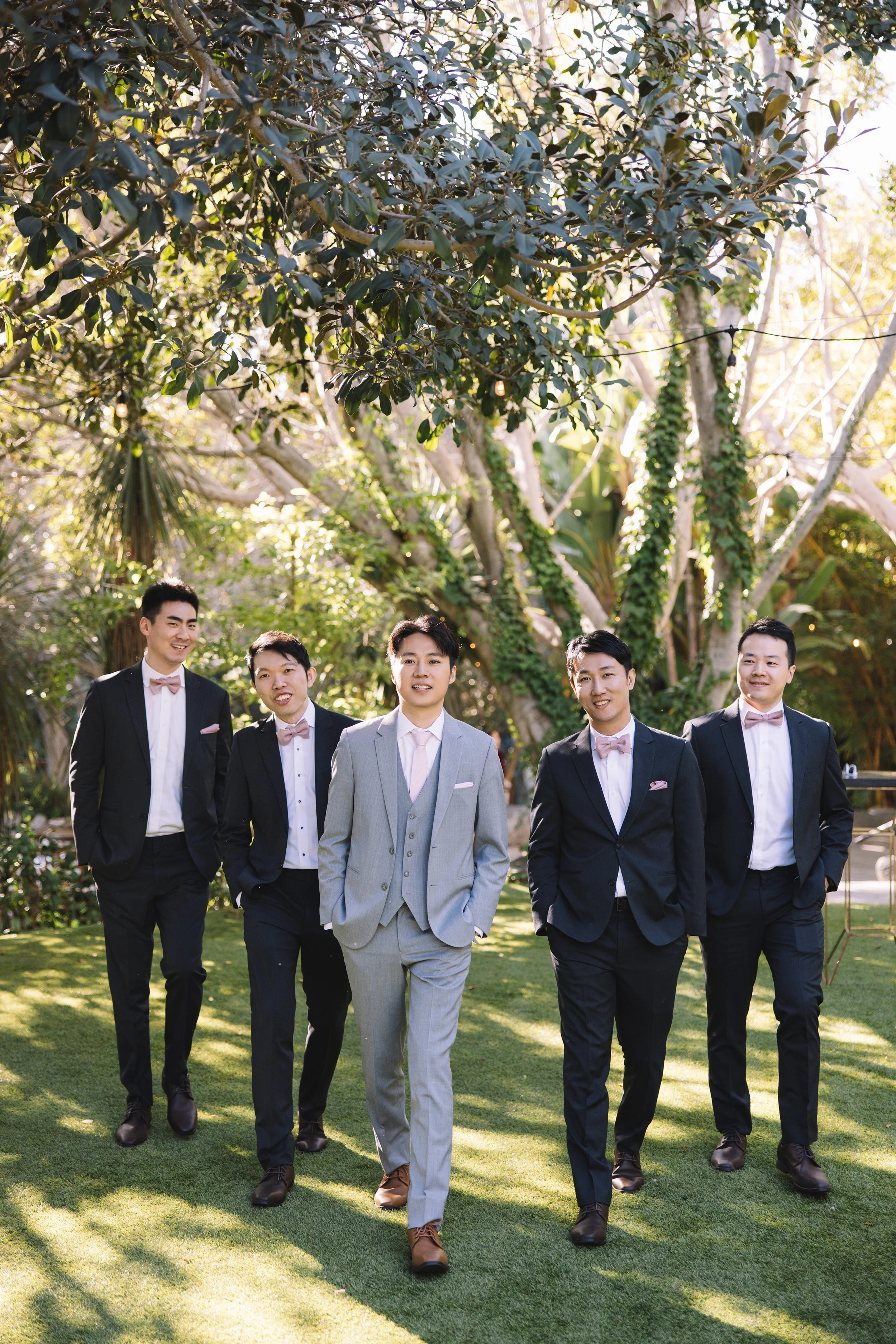 A group of five men dressed in suits walking outdoors in a garden with trees and greenery.