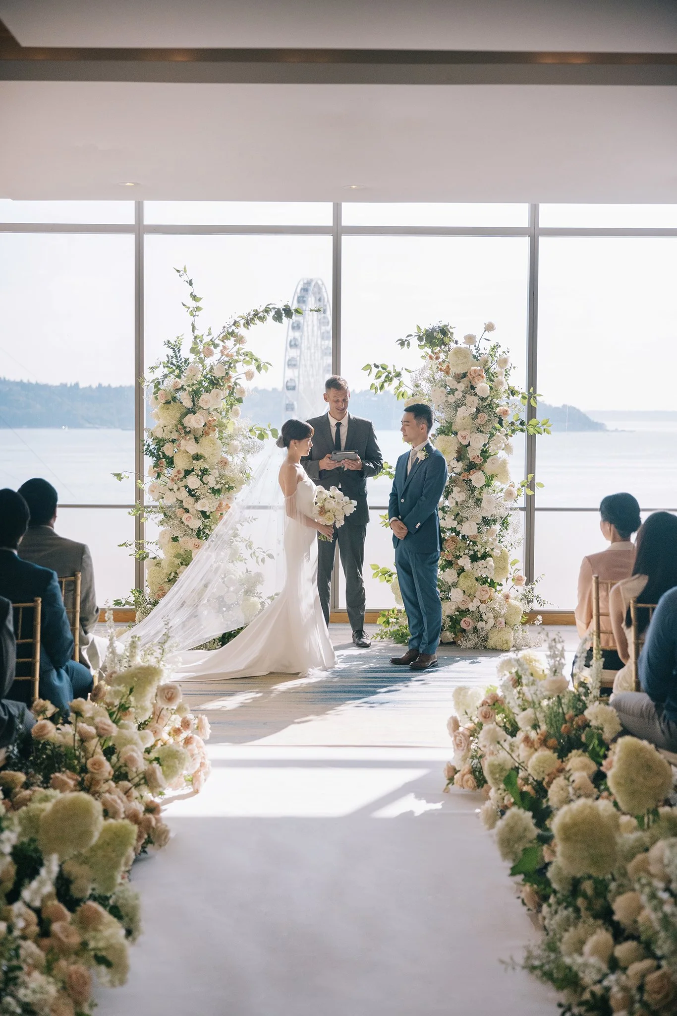 A wedding ceremony taking place indoors near large windows overlooking a body of water and a ferris wheel. The bride and groom stand facing each other, with the officiant in between, as guests sit on either side. The scene is decorated with white and