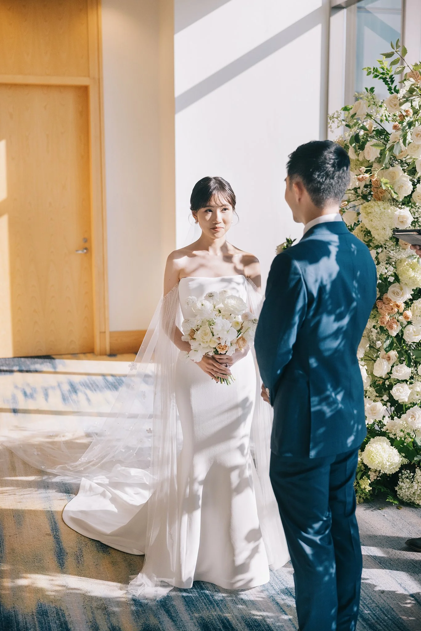 A bride and groom exchanging vows in a bright room with sunlight and a bouquet of white flowers.