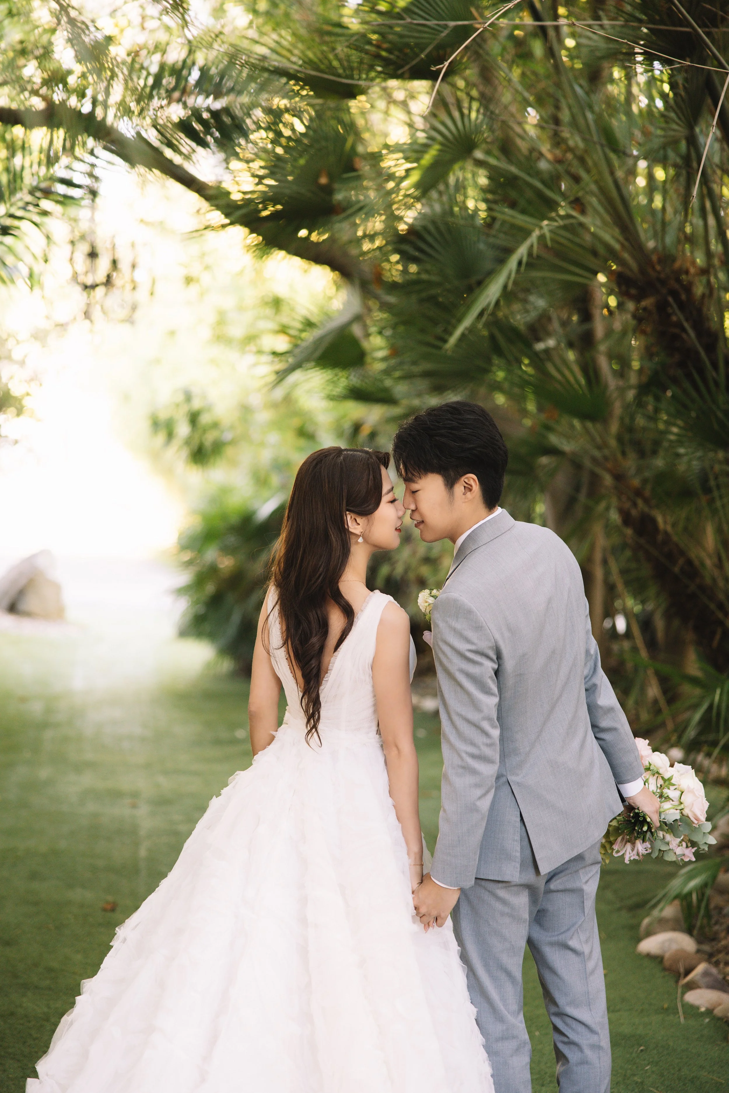 Bride and groom facing each other, holding hands, in a lush green garden.