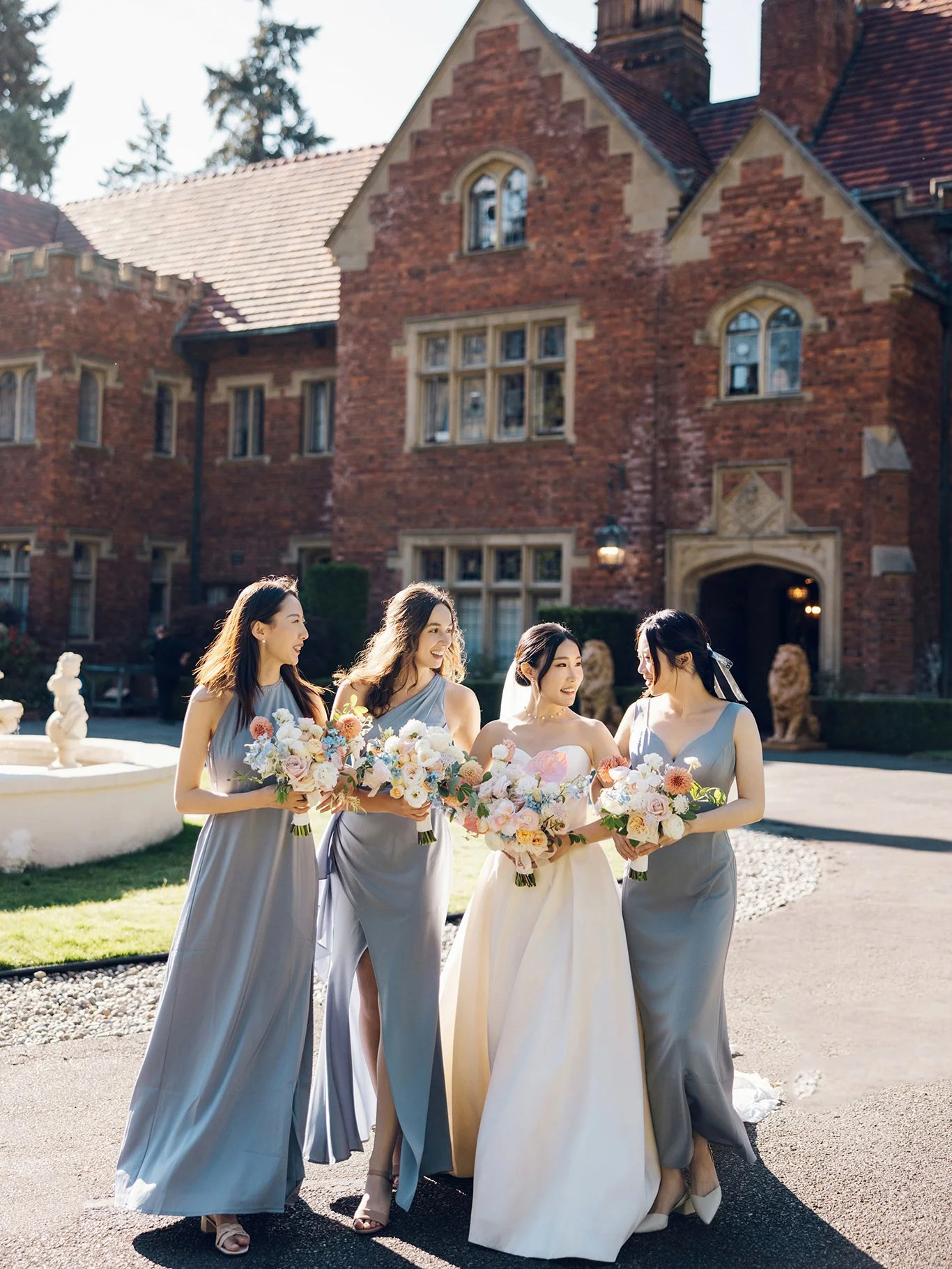 A bride and three bridesmaids smiling and holding bouquets in front of a large brick estate. The bride wears a white strapless gown with a veil, and the bridesmaids wear matching gray dresses with high slits.