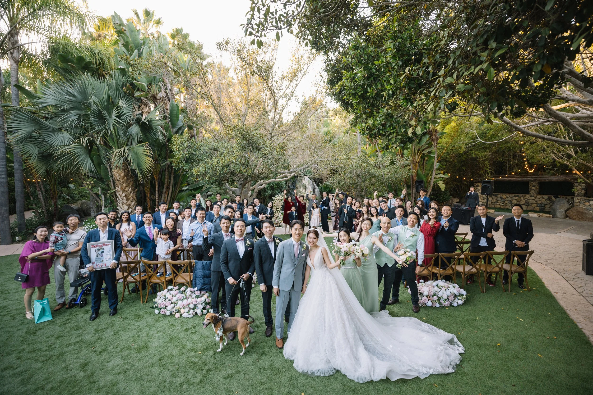 A large wedding party gathered outdoors in a lush garden setting with green trees and plants. The bride and groom are in the foreground, smiling and holding bouquets, surrounded by family, friends, and children. Some guests are standing, others sitti