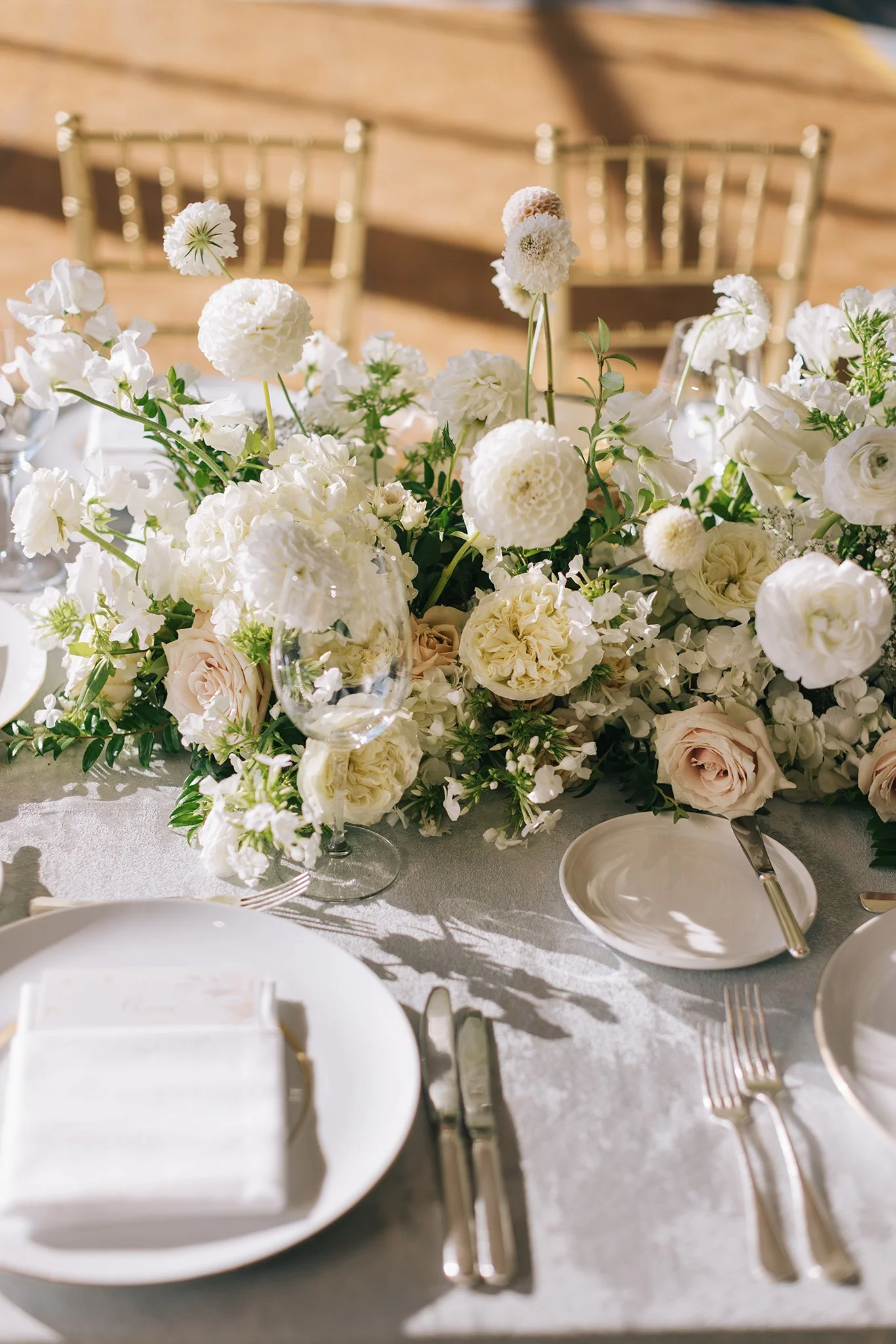 Elegant table setting with a lush white floral centerpiece including roses and dahlias, set for a formal event with white plates, glassware, silverware, and a napkin, on a light-colored tablecloth in a sunlit room.